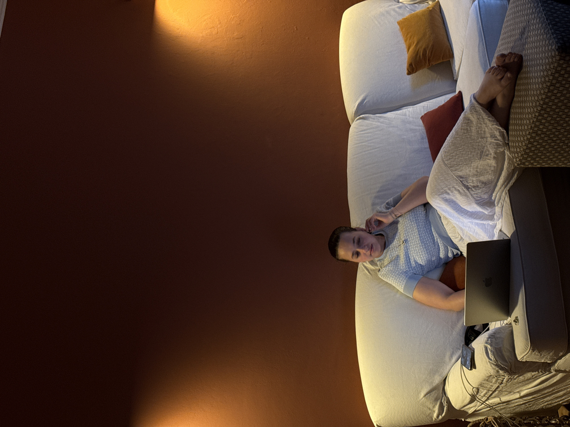 A person is lying on a light-colored couch with a laptop and a smartphone nearby. They are partially covered by a white blanket. The couch has several pillows, including one brown pillow, one orange pillow, and one patterned pillow at the end where the person's bare feet rest. The background features a dark, solid-colored wall, likely brown or dark orange, illuminated by a warm, indirect light source. The image does not contain enough information to determine if the location is in the city of Milan, Italy.