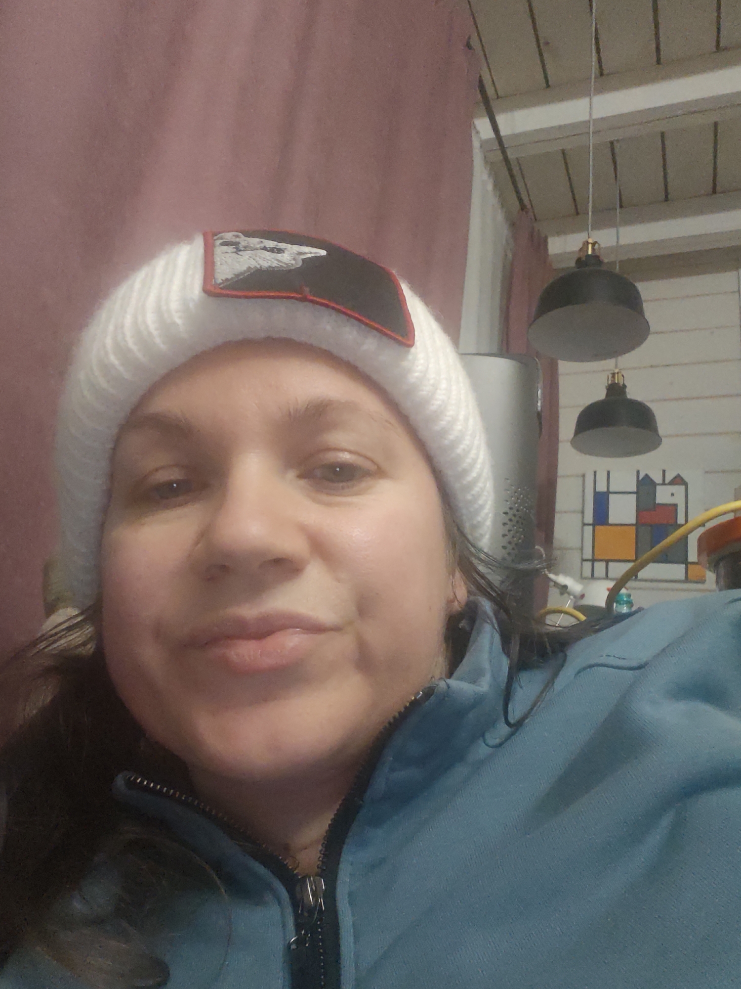 A selfie of a person with fair skin, wearing a white knitted hat with a patch featuring a bird (possibly an eagle) on a black background with a red border. The person also wears a blue zip-up jacket. Behind the person, there is a pink curtain, a white ceiling with exposed beams, two hanging black pendant lights, and a framed abstract painting of geometric shapes in primary colors and gray. The location cannot be confirmed as the city of Свистуха (кузяевский С/о), Russia, from the image alone.