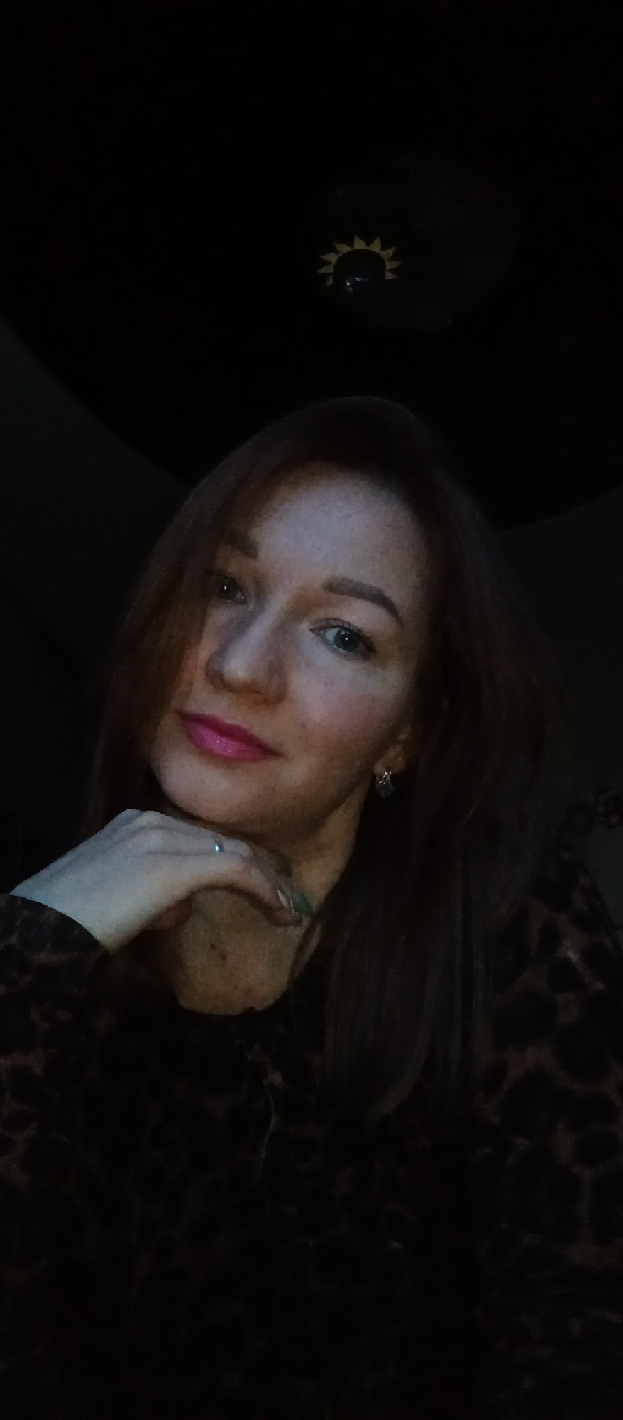 A dimly lit indoor portrait of a woman with reddish-brown hair, fair skin, and freckles. She has blue eyes and is wearing pink lipstick. Her right hand is supporting her chin. She is wearing a dark top with an animal print. A pair of earrings are visible on her left ear. In the background, a decorative light fixture resembling a sun is visible on the ceiling. The location of this image is unknown.