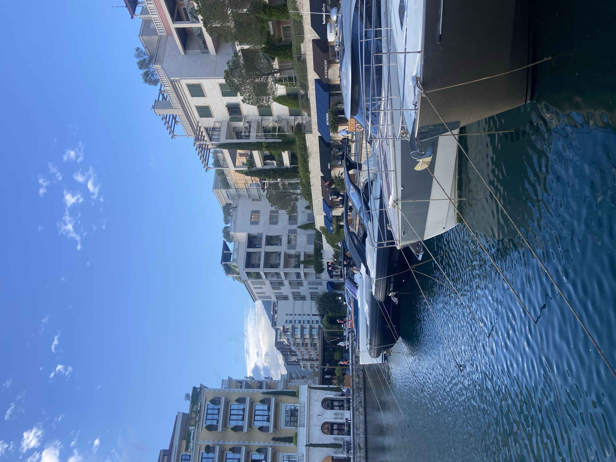The scene depicts a marina in Tivat, Montenegro, featuring yachts docked along a waterfront promenade. The foreground is dominated by the side of a large, modern yacht, displaying mooring lines connecting it to the dock. The water of the marina is visible, with reflections of the yachts and buildings. The background consists of a row of upscale buildings with architectural styles that include balconies, arched windows, and decorative landscaping. Storefronts with awnings, including one labeled "GIVENCHY," line the promenade. People are seen walking or seated at outdoor cafes along the promenade. The sky above is blue with scattered clouds.