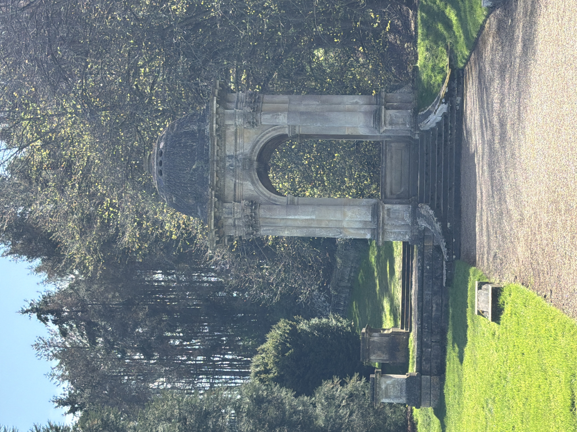 The image depicts a stone pavilion or folly with a domed roof, located in a garden setting, likely within the Malvern Hills area of the United Kingdom. The structure is built of light-colored stone, with an arched opening and decorative details. It is positioned at the top of a set of stone steps leading down into the garden. The scene includes trees in the background and manicured lawns. The photograph appears to be taken on a sunny day. There are no people visible in the image.