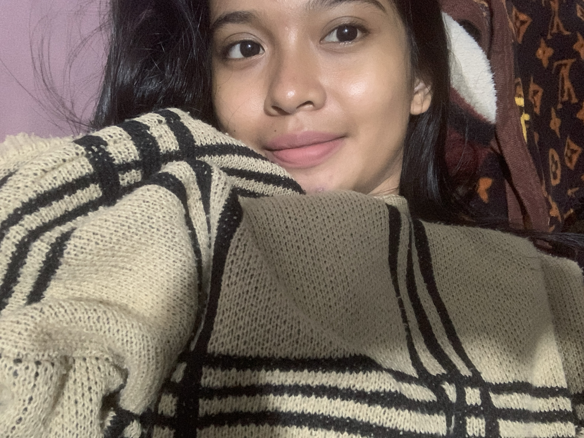 The image is a selfie of a young woman with dark hair. She has light to medium skin tone, dark eyes, and light pink lips. She's wearing a beige sweater with a black grid pattern. There's a patterned blanket behind her to the right. The background wall appears to be a light purple color. The image location cannot be determined to be Sungai Meriam, Indonesia, based on the visual information alone.