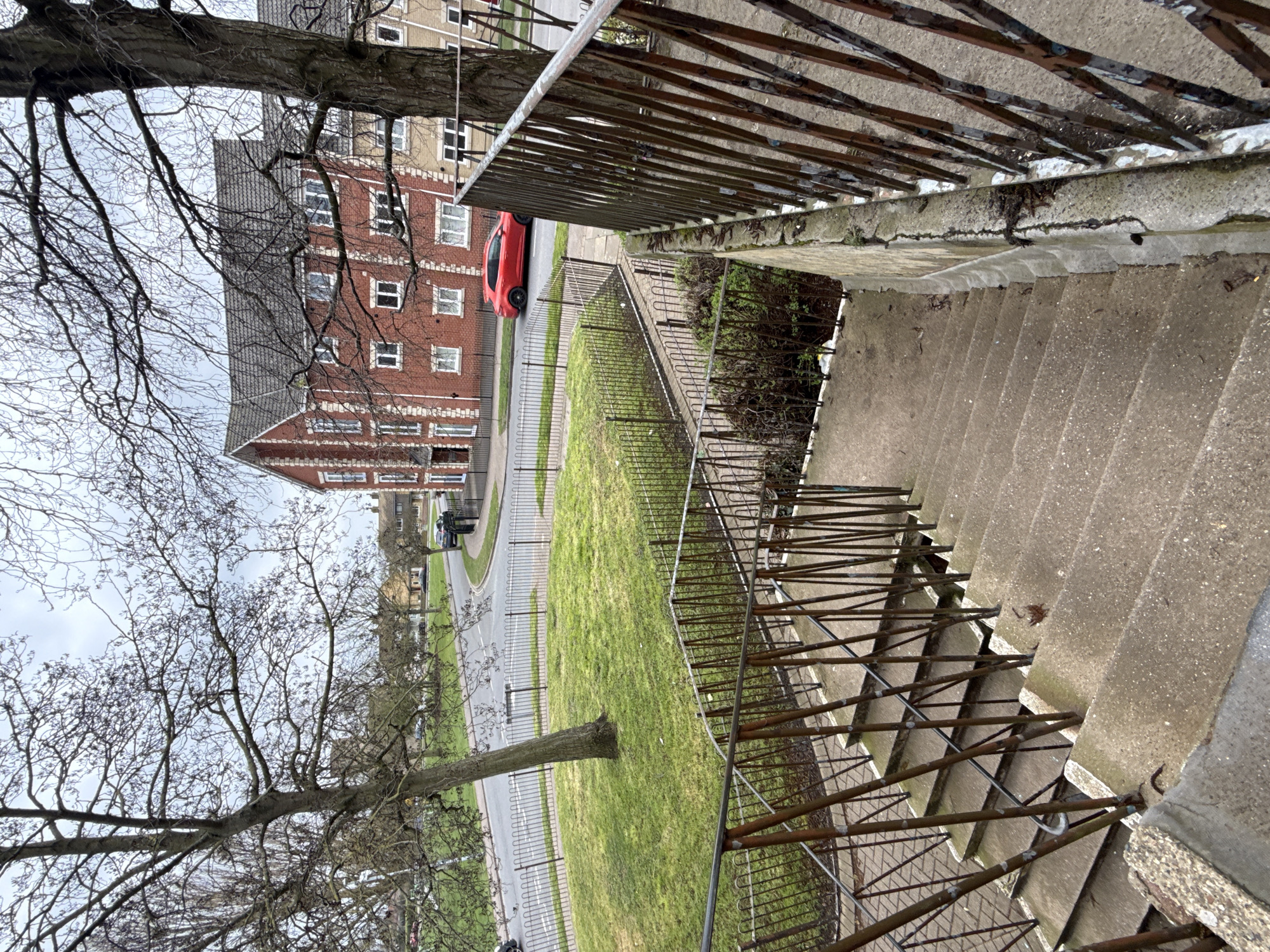 The image depicts a view from a concrete staircase with a rusted metal railing in Great Yarmouth, United Kingdom. The camera is positioned at the top of the stairs, looking down and outwards. Beyond the staircase and railing is a grassy area and a road. A red car is parked beside a red brick apartment building, which has multiple windows visible. A large bare tree is visible on the left of the image. The sky is overcast. There are no people visible in the image.