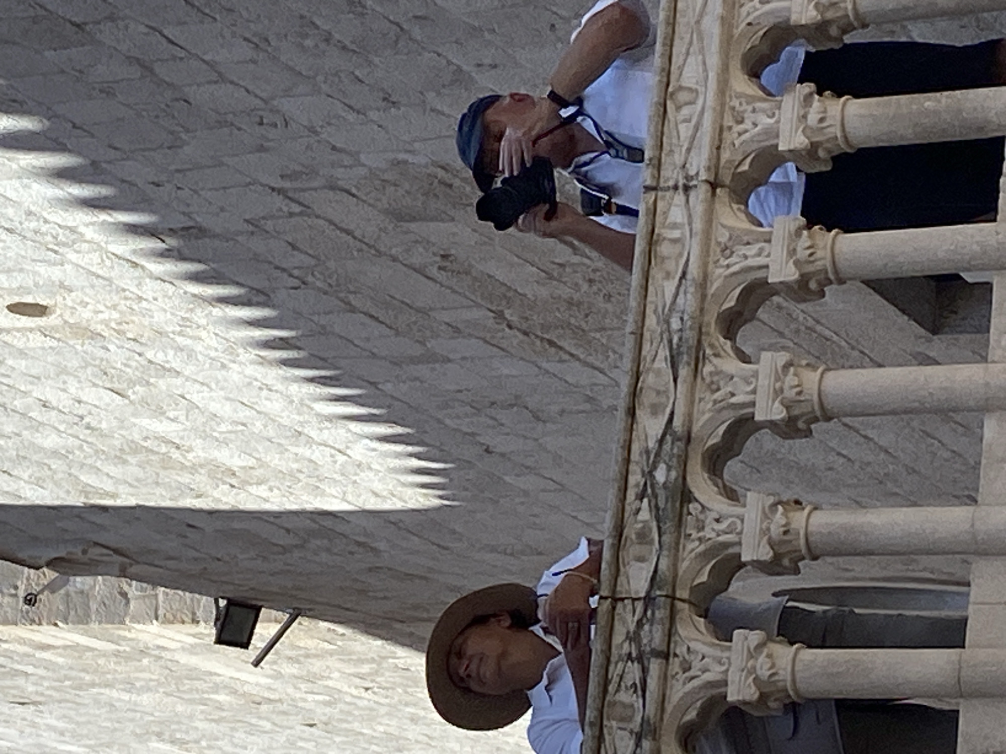 The image shows two individuals on a balcony with stone architecture. The person on the left is wearing a wide-brimmed hat and a white shirt, looking upwards. The person on the right is wearing a cap and a white shirt and is holding a black camera to their face, appearing to take a photograph. The balcony features ornate stone columns and arches, indicative of historical architecture. In the background, a stone wall is visible, with shadows cast across it from an unseen source, possibly foliage or architectural elements. One hint image shows a light fixture attached to the stone wall. The architectural style and the material of the wall suggest the location is within the historic city of Dubrovnik, Croatia, likely along the city walls or a similar ancient structure.