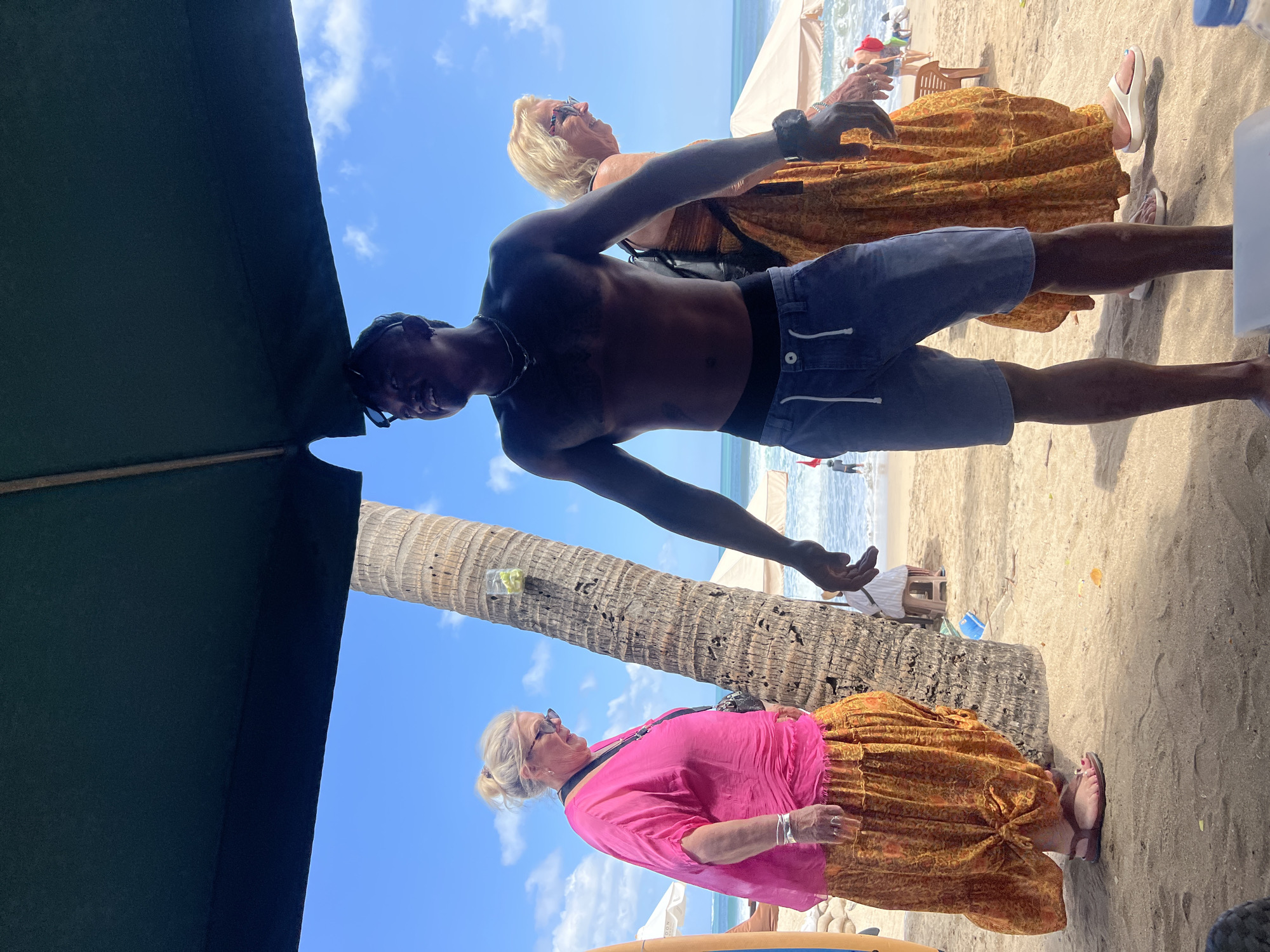 A man, shirtless and wearing blue denim shorts, stands on a beach in Kuta, Indonesia. He is looking to his left. To his left, a woman with blonde hair, wearing a bright pink top and an orange patterned skirt, is also looking to her left. Behind the man, another woman with blonde hair and wearing a similar orange patterned dress is visible, facing to her right. A palm tree is located between the man and the first woman. The background shows a sandy beach, the ocean with gentle waves, and a clear blue sky with scattered clouds. Beach umbrellas and chairs are visible in the distance. The scene appears to be under the shade of a large green umbrella.