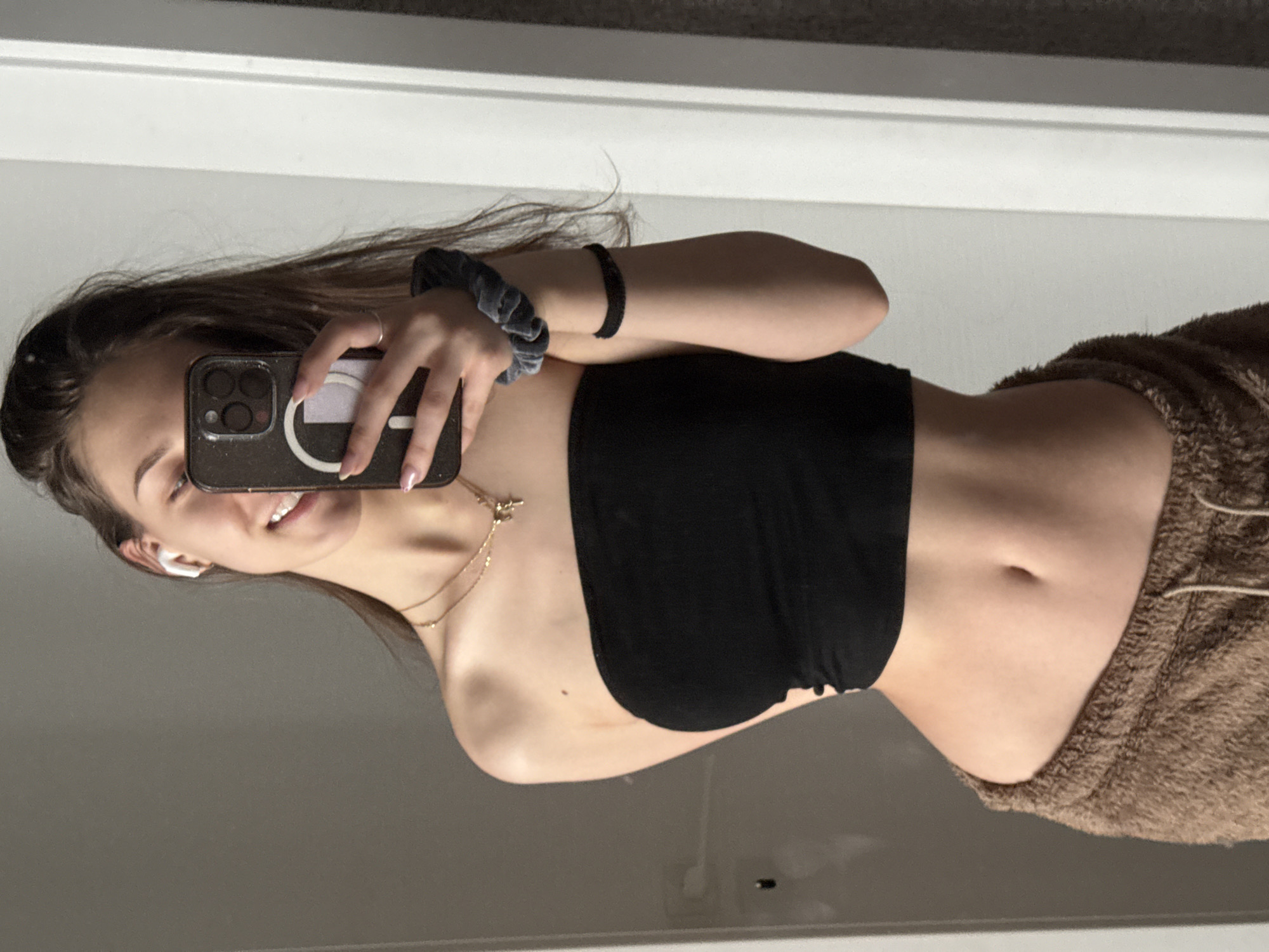 A young woman takes a selfie in front of a mirror. She is wearing a black tube top and brown pants. She holds a phone in her left hand. She is wearing a wireless earbud in her left ear, a scrunchie, and a bracelet on her left wrist, and a necklace. The location is not discernable from the image.