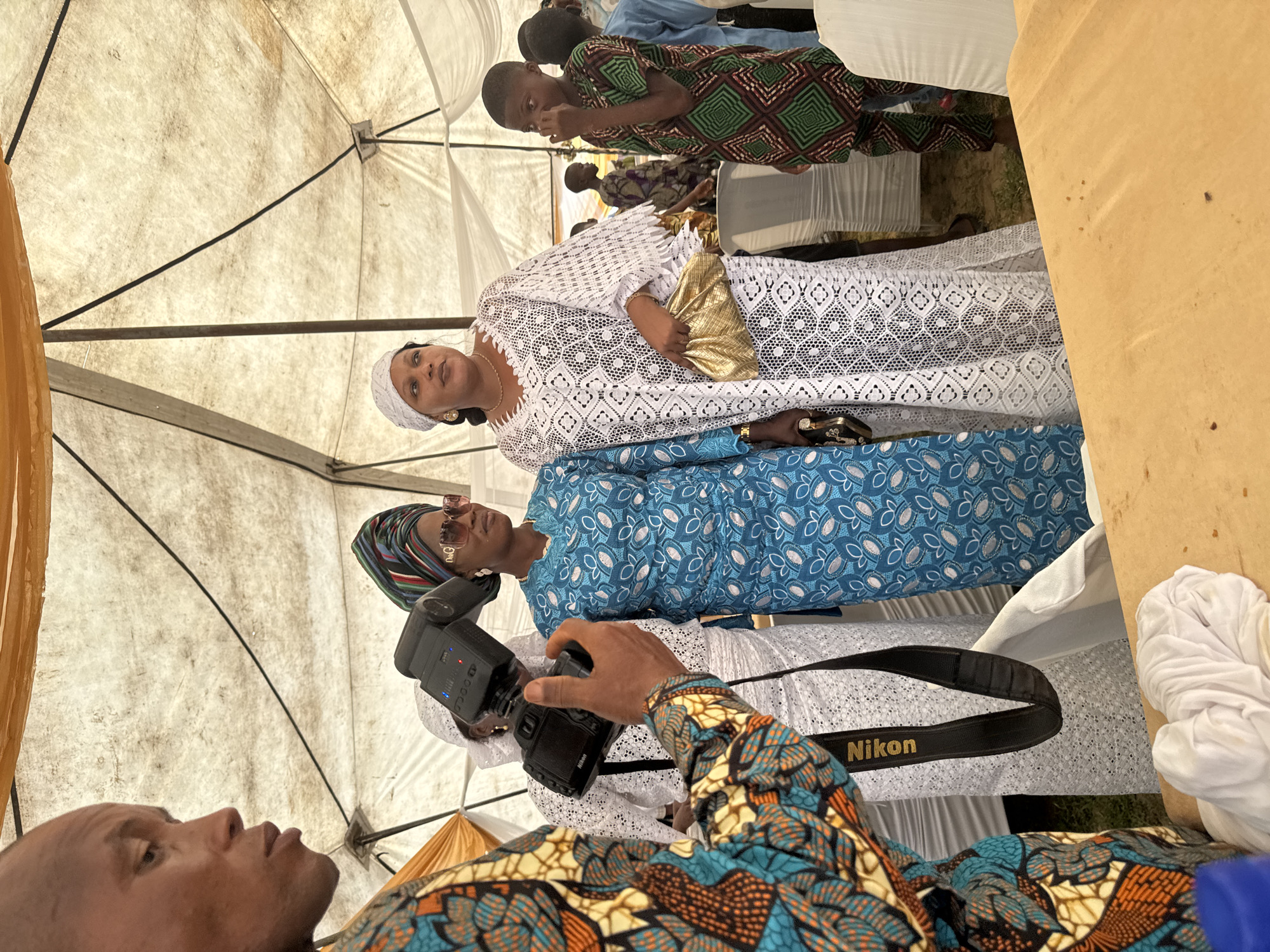 The image captures an outdoor event, possibly a ceremony, taking place under a large tent in Badagry, Nigeria. A man in a vibrantly patterned shirt is in the foreground, holding a Nikon camera with a flash attached, appearing to take a photograph. In the middle ground, two women are the primary subjects. The woman on the left wears a blue dress with a white pattern and a colorful headwrap, along with sunglasses and jewelry. The woman on the right is dressed in a white, intricately patterned lace dress and a white headscarf, carrying a gold clutch bag. Behind them, several other individuals, including children, are visible in the background, some of whom are also wearing traditional attire. The environment includes white draped fabric forming the tent structure and a wooden surface in the foreground, likely a table. The overall scene suggests a festive gathering in a community setting.