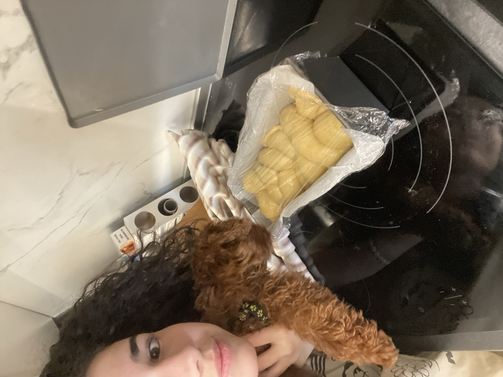 The image shows a selfie taken indoors, likely in a kitchen. A woman with dark, curly hair is visible in the foreground. She is holding a small, brown poodle with a yellow and green patterned collar. Behind them, on a countertop, there's a roll of aluminum foil and what appears to be challah bread inside a plastic bag. The countertop is marble. To the right is a black stovetop. An oven is located at the top left. There are no indicators of the city of Sofia, Bulgaria.