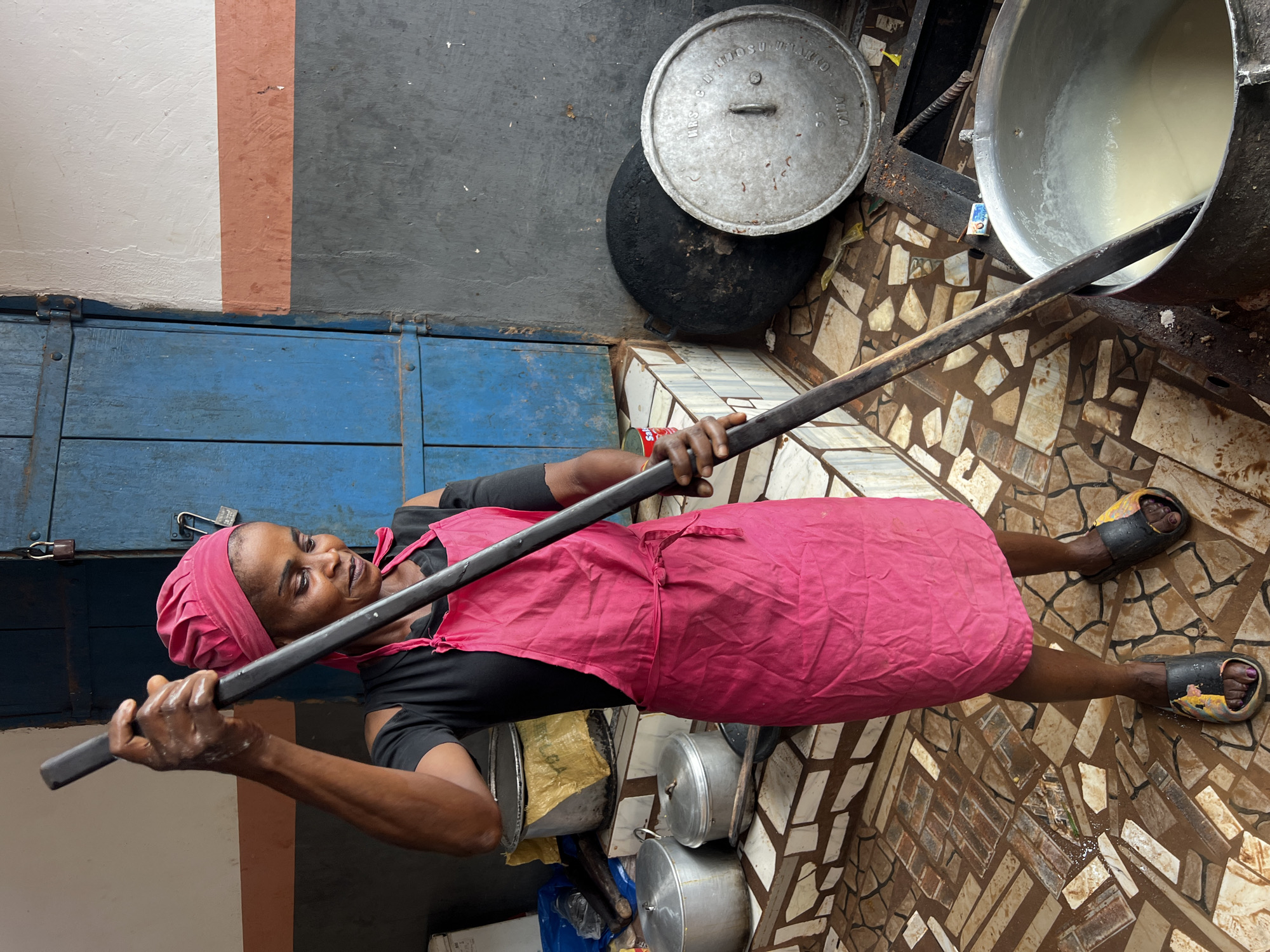In Nkpor, Nigeria, a woman wearing a pink headscarf and apron stands by a large pot, stirring its contents with a long wooden paddle. She is dressed in a black t-shirt with cold-shoulder cutouts. The scene appears to be an outdoor cooking area, indicated by the tiled floor, a blue wooden door with padlocks in the background, and various cooking implements such as pots and pans scattered around. A large, dark, round pot is visible to the right of the woman, alongside a silver pot with a lid. The woman's hands are wet, suggesting she is actively engaged in cooking. The overall environment appears to be a functional, possibly commercial, food preparation space.
