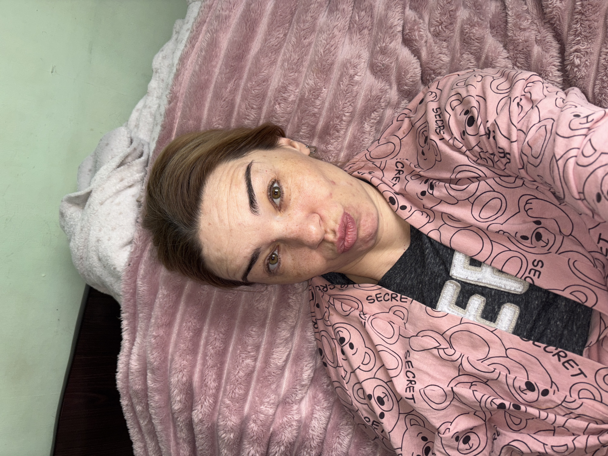 A woman is lying on a pink blanket, looking at the camera. She is wearing a pink robe with a teddy bear pattern and a grey t-shirt with the letters "EF" on it. The background is a light green wall.