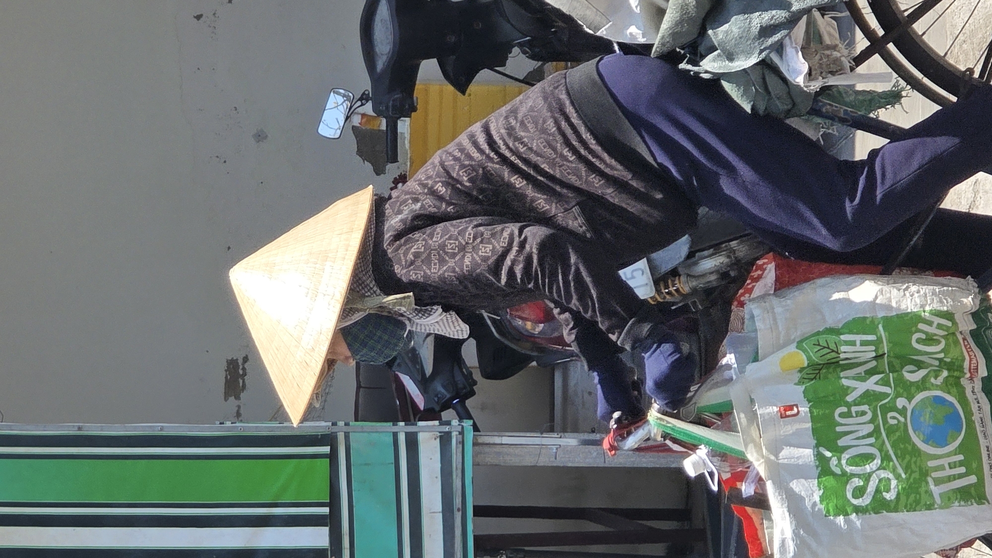 A person wearing a conical hat and patterned clothing is bending over, possibly working on something near a motorcycle heavily loaded with various items, including bags with green lettering that says "SỐNG XANH". A blue and green striped object is visible to the left. The location is likely in Bắc Nha Trang, Vietnam.