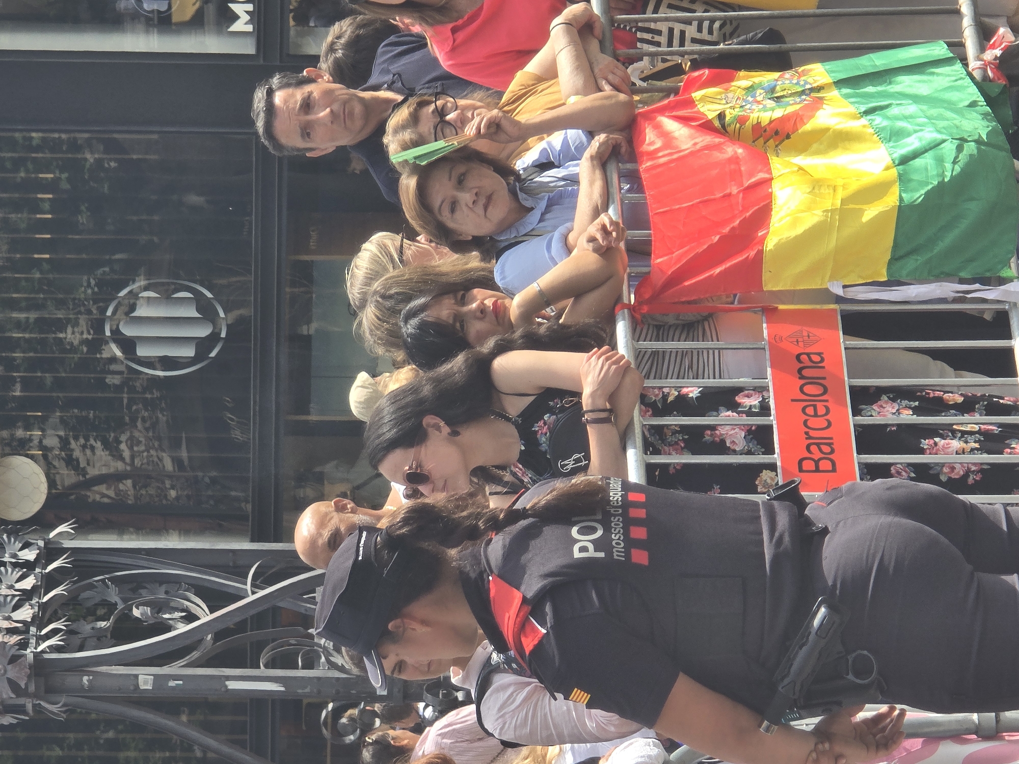 This image captures a scene from what appears to be a public event in Barcelona, Spain. The primary subjects are a group of people, both civilians and law enforcement, gathered behind a metal barrier. In the foreground, a woman wearing a police uniform is visible, with "POL" and "mossos d'esquadra" clearly printed on her vest, along with red squares. She has a firearm holstered on her hip.
The civilians in the background are diverse, with varying ages and expressions. Some are looking towards something off-camera, suggesting they are spectators. One person is holding a green fan, and another is holding a flag with red, yellow, and green stripes, and a coat of arms, which could be a national or regional flag. The flag is draped over the barrier.
The setting appears to be outdoors, with sunlight suggesting it is daytime. The background shows elements of a building with glass windows, and a decorative metal fence or gate in the lower part of the image. A bright red sign with white text reading "Barcelona" is attached to the barrier.
The emotions of the individuals are difficult to discern definitively due to the image's orientation and resolution, but the overall atmosphere seems to be one of engagement with an event. The presence of police and a gathered crowd indicates a public gathering, possibly a parade, protest, or festival.