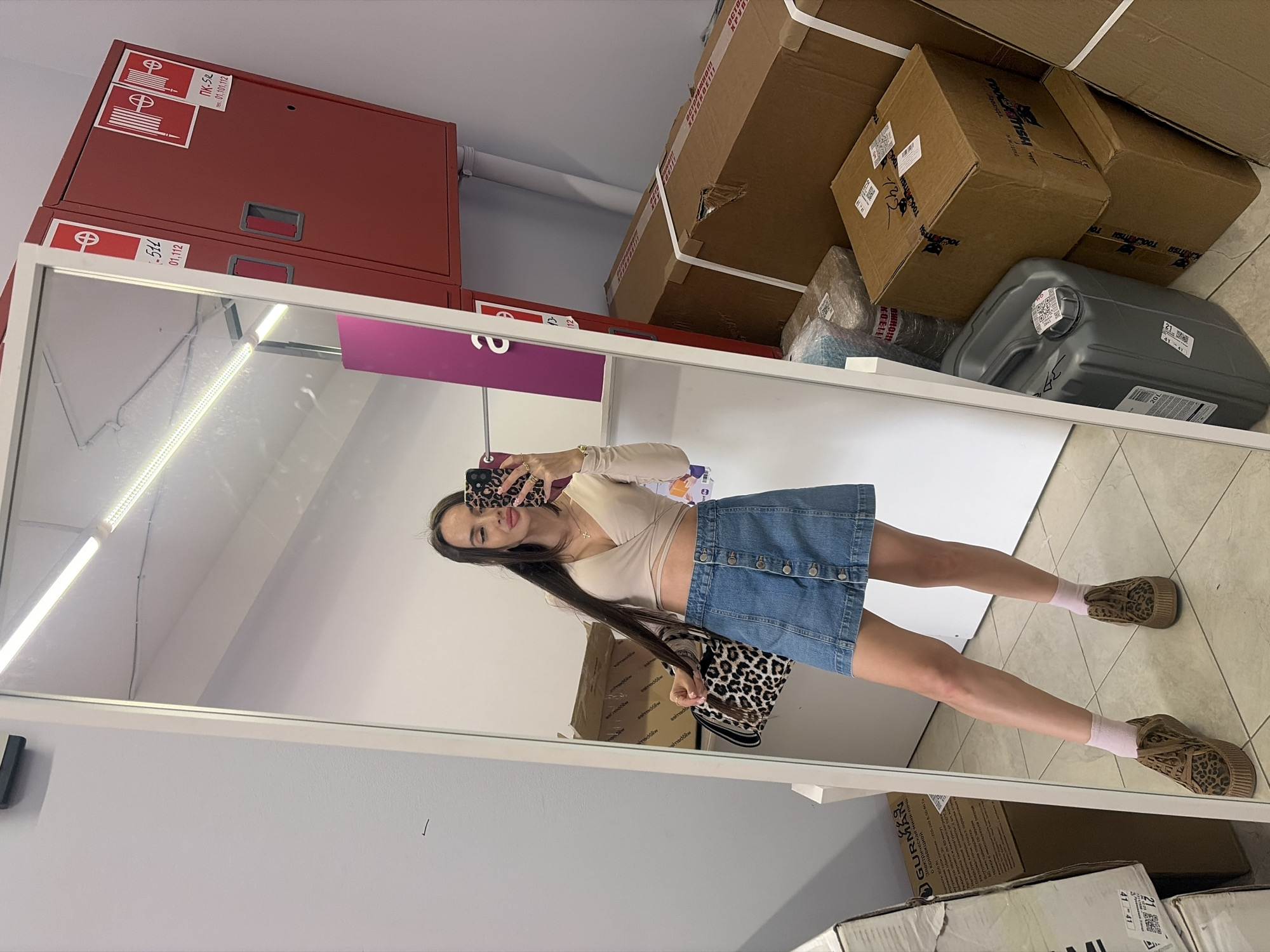 The image displays a mirror selfie of a young adult female taken in an indoor commercial or storage facility located in Saint Petersburg, Russia. The female, characterized by long dark hair, is wearing a light beige long-sleeved wrap-around crop top, a button-front denim mini-skirt, light pink ankle socks, and leopard-print platform sneakers. She holds a leopard-print shoulder bag in her left hand, which also clutches a section of her hair. Her right hand holds a smartphone with a leopard-print case, positioned for the selfie, with her lips slightly puckered. A delicate gold cross necklace is visible.
The environment reflected in the mirror features light grey walls and a light-colored tiled floor. Overhead fluorescent strip lights are visible in the reflection. To the left of the subject, a large cardboard box is partially visible, bearing the Cyrillic text "GURMAN" and "НЕ ВЕРТИ!" (DO NOT TWIST!). To the right of the subject, two red fire hose cabinets are mounted on the wall. The left cabinet is labeled "ПК-51L / 01.112" and has a sticker indicating "ТЕХ. ОБСЛУЖИВАНИЕ 01.01.2023" (Technical Service 01.01.2023). The right cabinet is labeled "ПК-52 / 01.10.112". A partially visible purple sign with "а" and "13-12" is seen between the subject and the fire cabinets. Below the fire cabinets and extending to the right, numerous cardboard boxes are stacked, some secured with white strapping. Visible text on these boxes includes "ОСТОРОЖНО ХРУПКОЕ!!" (CAUTION FRAGILE!!), "TOOlTECH" branding, and "Made in Russia". A grey 20-liter plastic canister with a QR code sticker is also visible on the floor among the stacked boxes.
