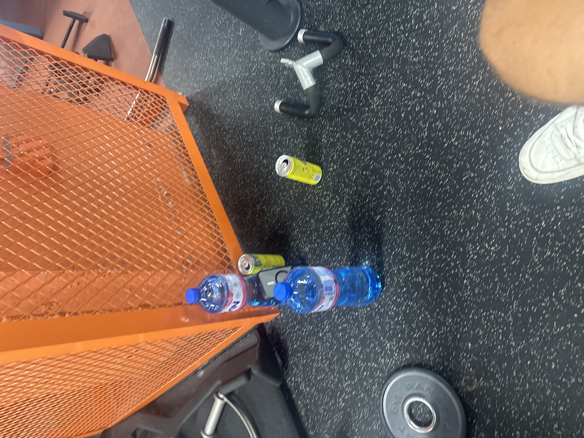 The image shows a scene inside a gym, likely located in Plovdiv, Bulgaria. The floor is covered in a dark gray speckled rubber material typical for gym flooring. Present are pieces of gym equipment: a leg press, and a weight plate. Two blue water bottles, and two yellow aluminum cans are lying on the floor. A person's leg wearing a white sneaker is visible at the edge of the frame.