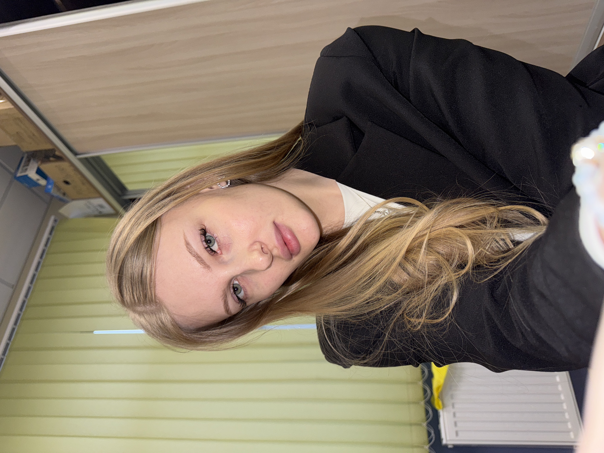 A young woman with blonde hair is posing for a selfie indoors. She is wearing a black blazer over a white top. She has light skin and blue eyes. The background appears to be an office or similar workspace with light-colored window blinds and a cabinet. No specific evidence suggests the location is in Perm, Russia.