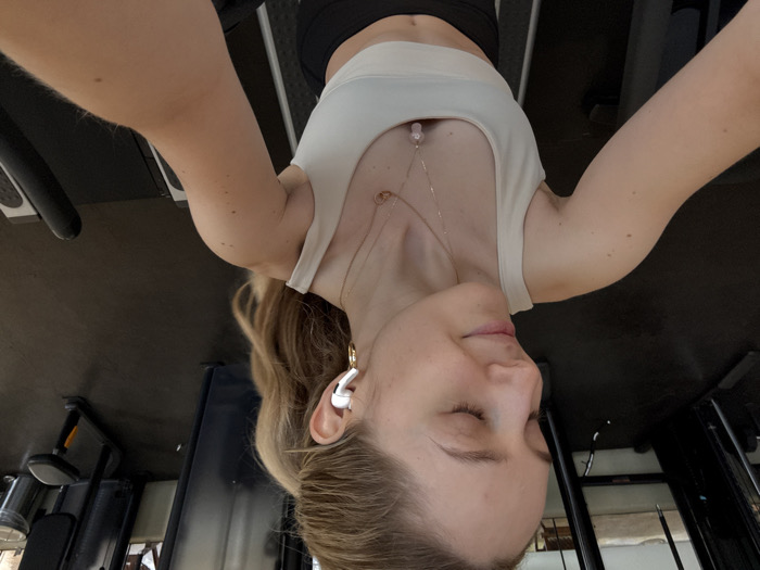 The image depicts a close-up, selfie-style shot of a young adult female located within an indoor gym facility in Mexico City, Mexico. The subject, possessing a light complexion and long blonde hair tied in a high ponytail, is shown from the chest up, with her head slightly tilted to the right and eyes closed, displaying a subtle smile. She is wearing a light-colored athletic crop top and dark-colored athletic bottoms. A white wireless earbud is visible in her right ear, along with a gold-colored hoop earring. Two layered gold-colored necklaces with pendants, one circular and one featuring a pinkish stone, adorn her neck. Her right arm is extended, holding the camera, with the top of a black treadmill handrail visible in the lower right foreground. The background is blurred, revealing dark gym flooring and indistinct black-framed gym equipment, possibly including weight machines and a silver-colored waste bin, suggesting a commercial fitness environment. The lighting is bright and even.