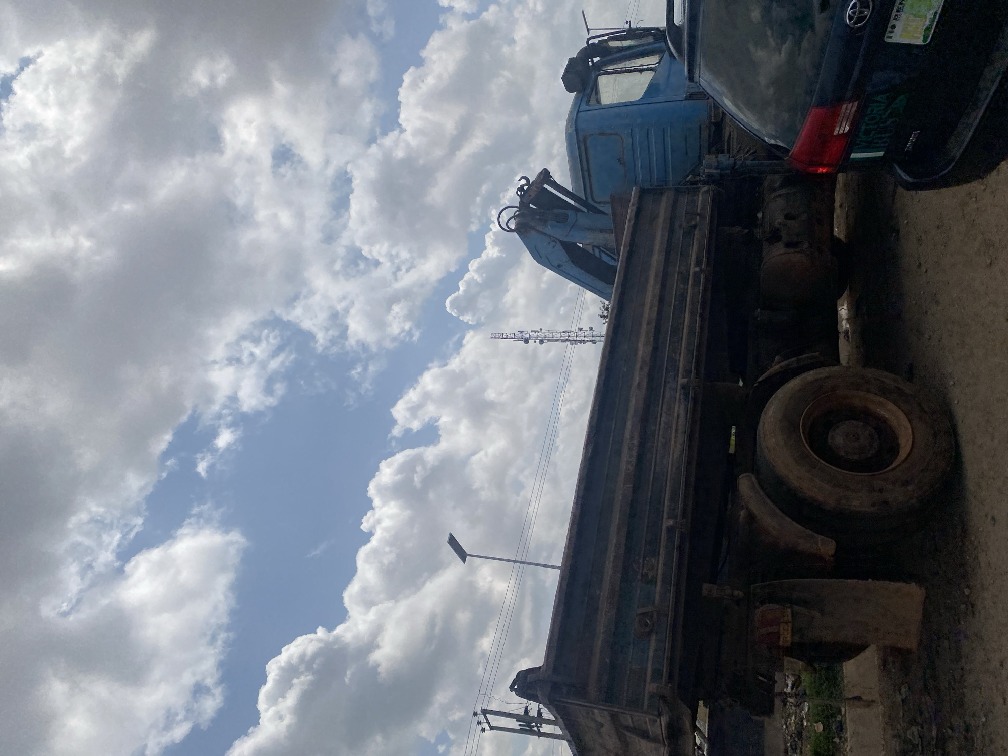 The image depicts a street scene in Ado, Nigeria, under a partly cloudy sky. In the foreground, a dark-colored SUV is parked on the right side of the frame. To its left and dominating the left side of the image is a large, blue dump truck with a crane attachment. The truck appears weathered and dirty. Its cargo bed is empty and rust is visible on its exterior. The rear tire and fender of the truck are prominent. The ground is unpaved and appears to be a dirt or gravel road. Behind the truck, a tall telecommunications tower is visible in the distance, as well as a street light on a pole. Power lines are also present in the background. The scene suggests a commercial or industrial area within the city. There are no people visible in the image.