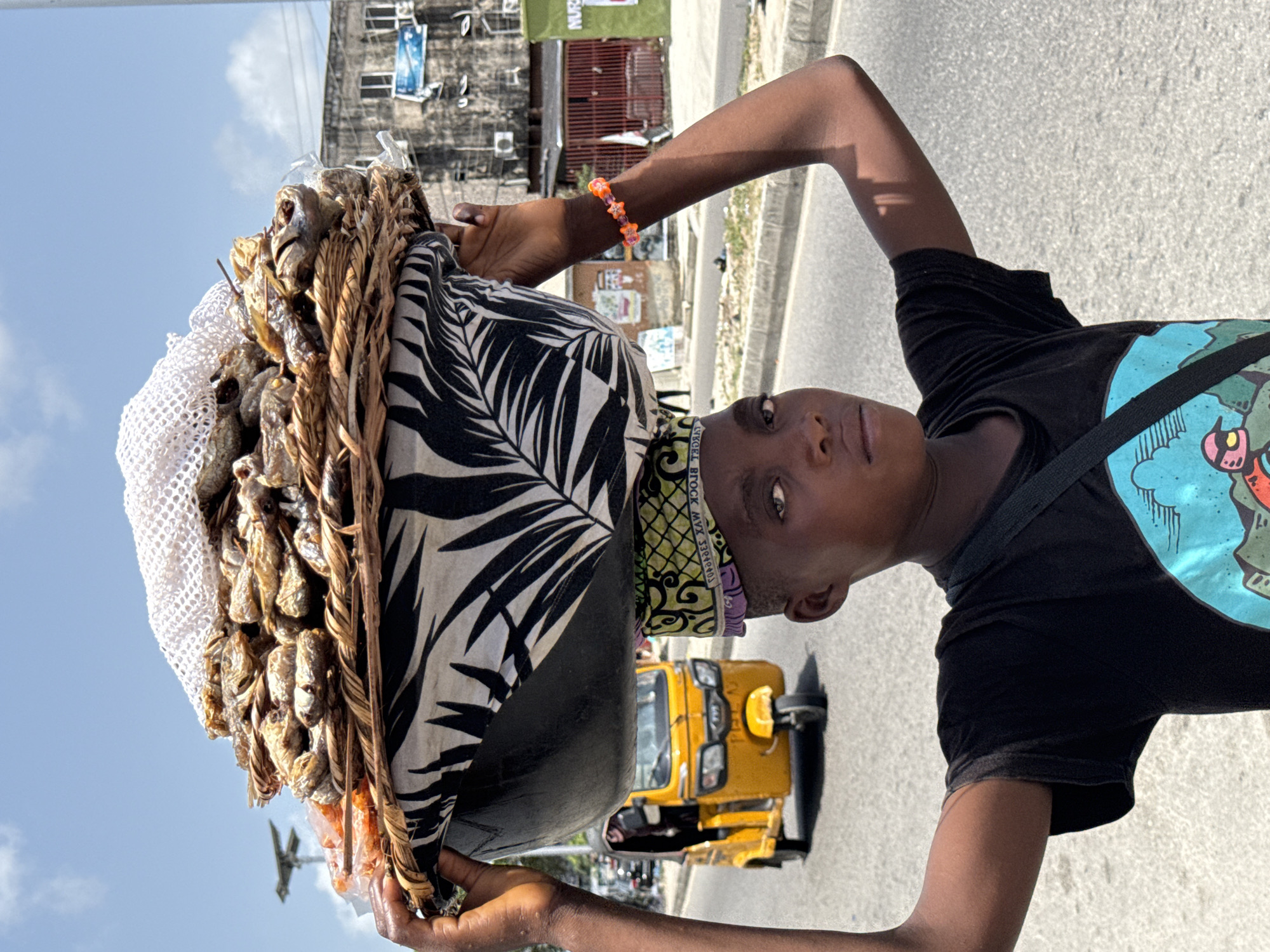 In Ado, Nigeria, a young person stands on a street, carrying a large basket on their head. The basket is filled with dried fish, wrapped in a white mesh netting and lined with a black and white patterned fabric. The person wears a black t-shirt with a colorful graphic design and a strap across their chest. They also have an orange beaded bracelet on their left wrist and a patterned head wrap. A yellow auto rickshaw (keke) is visible in the background on the asphalt road. Buildings, utility poles, and other vehicles are also visible. The sky is light blue.