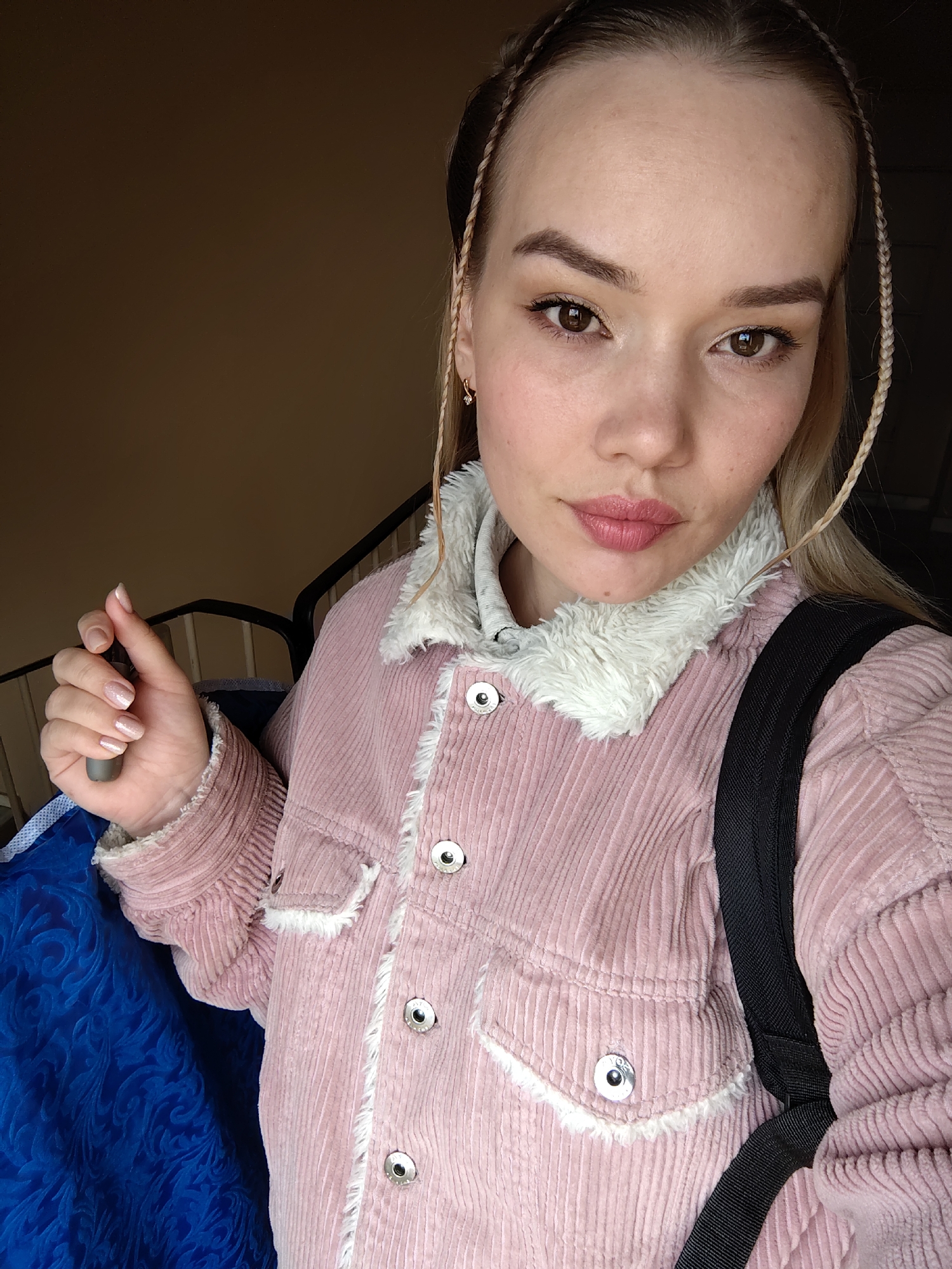 A young woman is captured in a close-up selfie, looking directly at the camera with a composed, slightly pouting expression. She has fair skin, prominent dark brown eyes, and light brown eyebrows, with a hint of subtle makeup including eyeliner and lip color. Her light blonde hair is styled with thin braids framing her face, and she wears small, gold-toned hoop earrings with clear stones.
She is dressed in a soft pink corduroy jacket featuring a white faux fur collar and matching faux fur trim along the seams and pocket flaps. The jacket is fastened with silver-toned buttons. A black backpack strap is visible over her right shoulder. In her left hand, she holds a small, dark grey cylindrical object, possibly a vape pen or a similar device. Her fingernails are neatly manicured with a light, shimmering polish.
To the left of the frame, a segment of a blue fabric with a raised, ornate pattern is visible, likely part of an item she is carrying or nearby. The background is simple, featuring a plain brown wall on the left and a black metal railing on the right, suggesting an indoor setting such as a hallway or stairwell. The lighting is soft and even, indicating a daytime shot. The image was taken in Марковский, Russia.