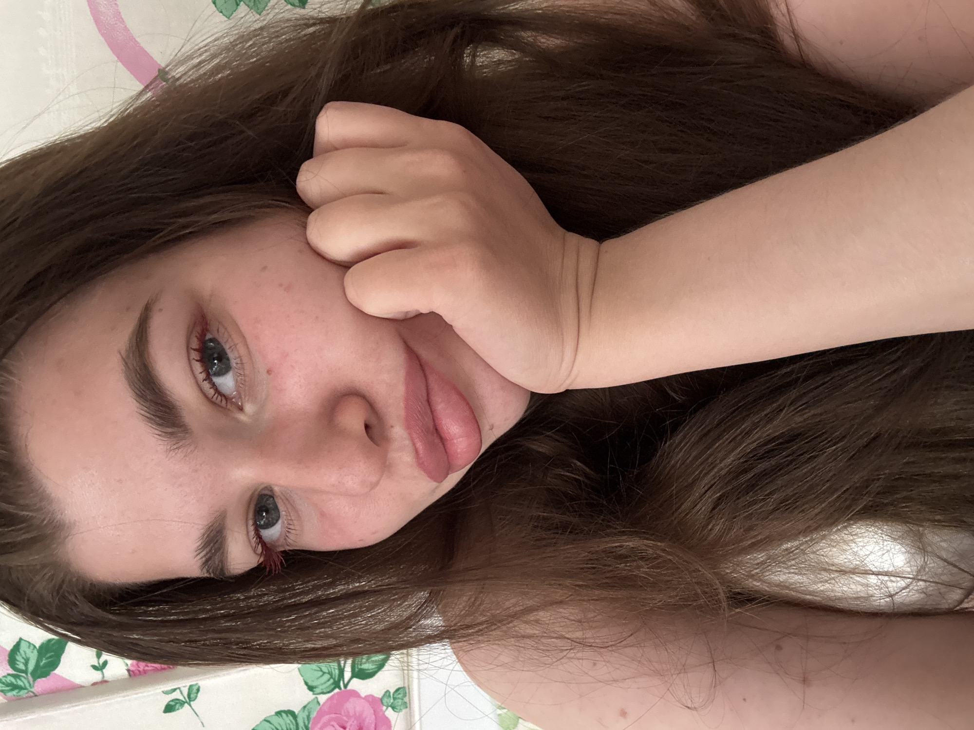 A young woman with long brown hair rests her head on a floral-patterned surface. Her hand is positioned near her face. She has fair skin, prominent eyebrows, and blue eyes with red eyelashes. Her lips are slightly parted, and she appears to be in a relaxed pose. There is no information about the city of Мещёры, Russia.