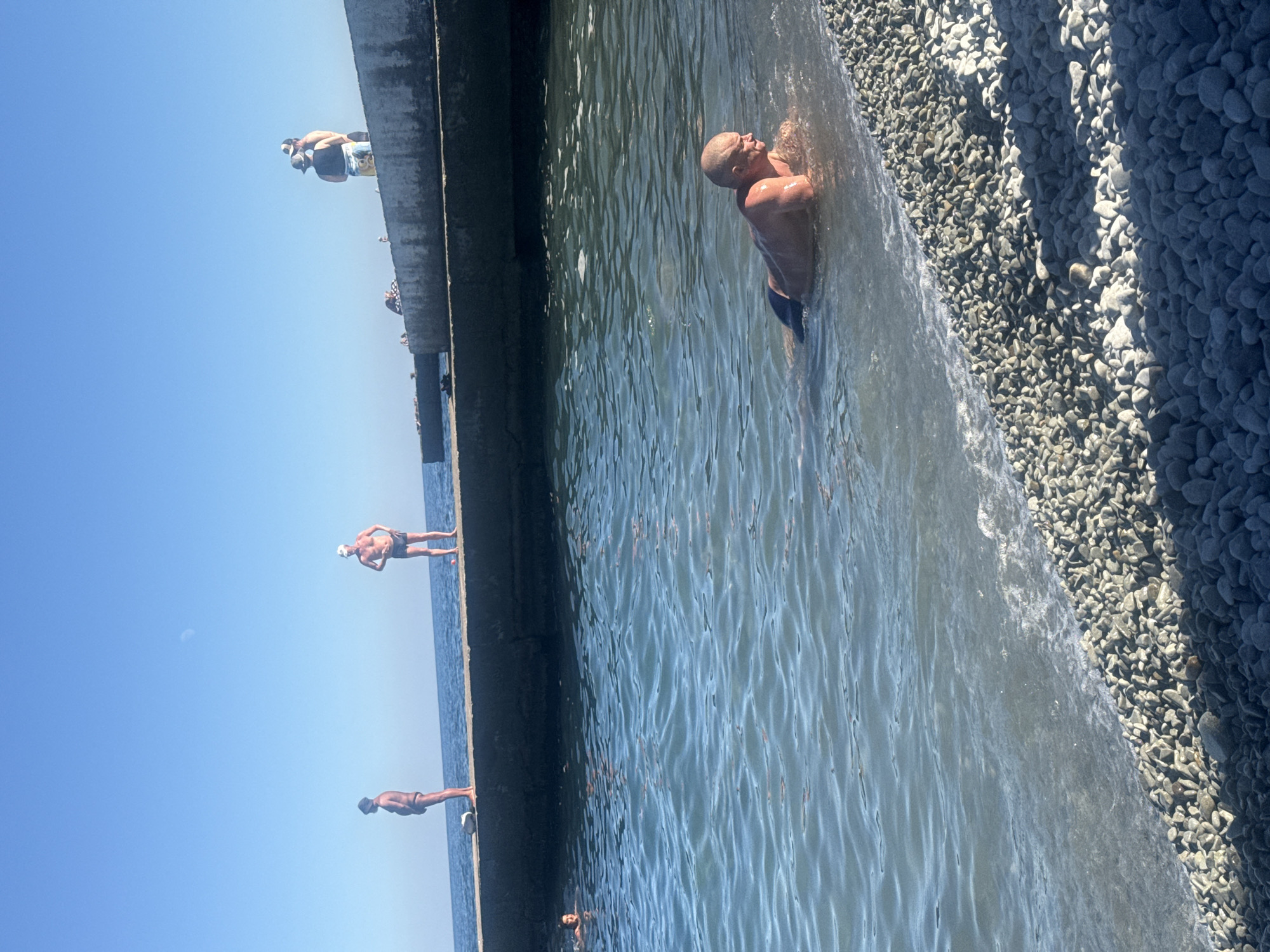 The image shows a sunny day at a beach with a clear blue sky. A concrete pier extends into the water, and several people are visible on it and in the water. In the foreground, a man with a shaved head is lying in the shallow water near the pebble beach, facing right. His upper body is out of the water, and he appears to be resting. Further in the water, a woman with dark hair is swimming and looking towards the camera. On the pier, there are several individuals, some standing and some appearing to be sitting. One man on the pier is standing with his hands on his hips, wearing a hat and dark shorts. Another man with a hat is standing next to him. Further down the pier, more people are visible, some in swimwear. The water is calm with gentle waves lapping at the shore, and the beach is covered in smooth, grey pebbles. The overall scene depicts a leisurely day at the seaside in Partenit, Ukraine.