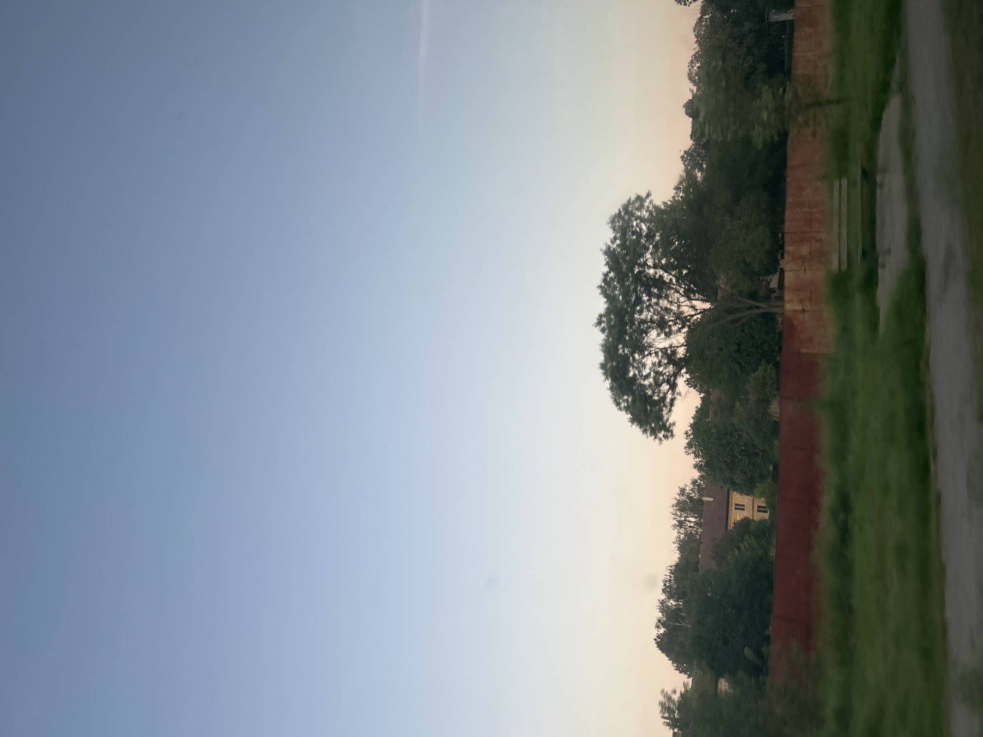 The image depicts a serene outdoor scene, likely taken at dusk or dawn, with a gradient of soft blue and peach hues in the sky. In the middle ground, a row of lush green trees forms a natural barrier, behind which a yellow building with a red roof is partially visible. A rust-colored wall or fence runs horizontally across the frame, separating the treeline from the foreground. In the foreground, a grassy area is visible, with a concrete path or walkway running through it. A weathered wooden bench with metal legs sits on a concrete slab, suggesting a public park or rest area. The overall impression is one of tranquility and natural beauty. This image may be located in Elin Pelin, Bulgaria, as indicated by the prompt.