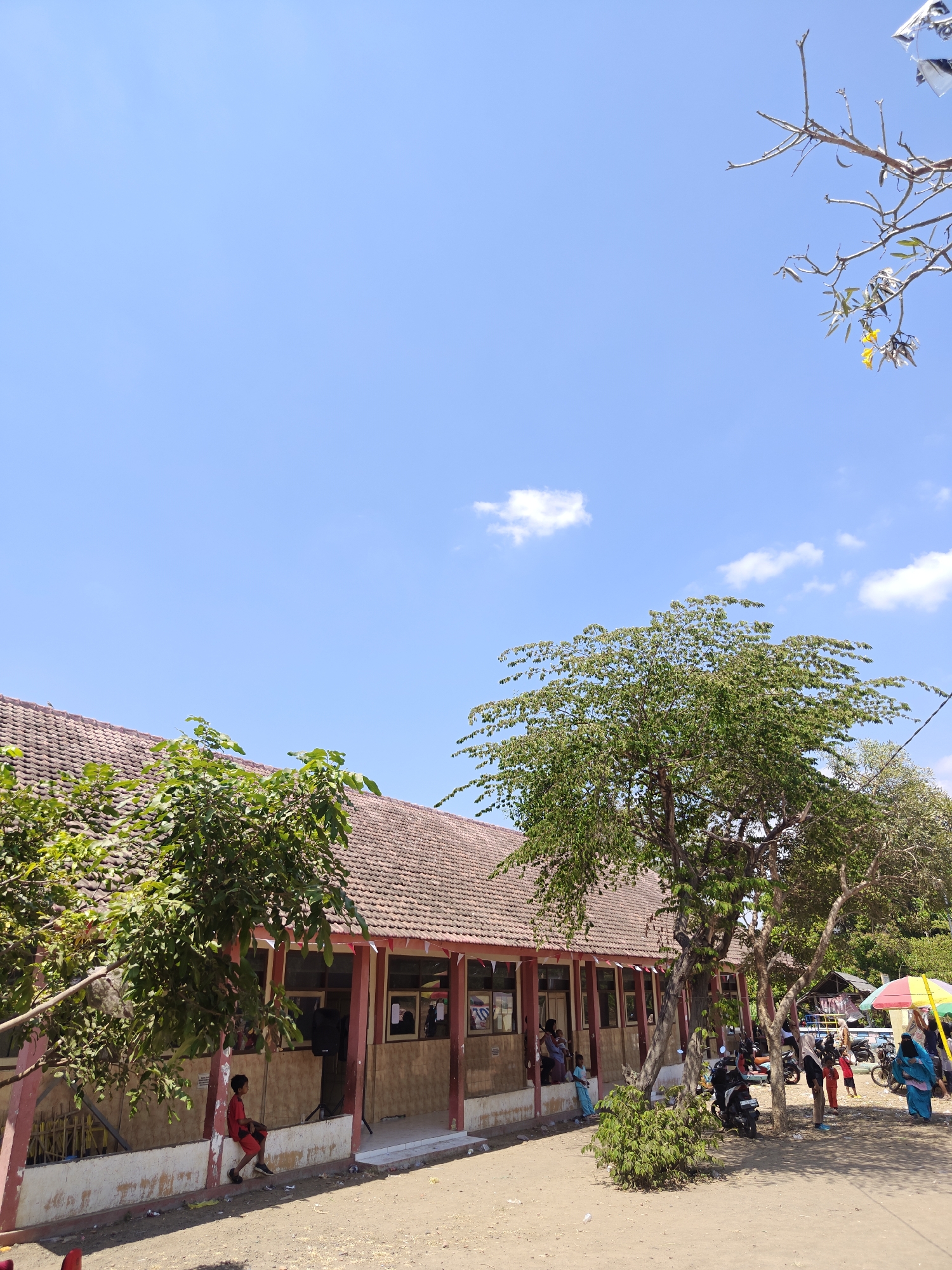 A low-angle, full shot depicts a one-story school building with a red tiled roof under a bright, clear blue sky in Cermee, Indonesia. The building has a covered walkway with red pillars and visible windows, some of which display framed pictures. In front of the building, the ground is sandy and dusty. Several people are visible, including a young boy in a red shirt and shorts sitting on a low wall, a woman and child standing near the entrance, and a group of people with motorcycles and a colorful umbrella in the background. Trees with green foliage are scattered around the building, one large tree prominently featured in the foreground. The overall scene suggests a warm, sunny day with some activity taking place outdoors.
