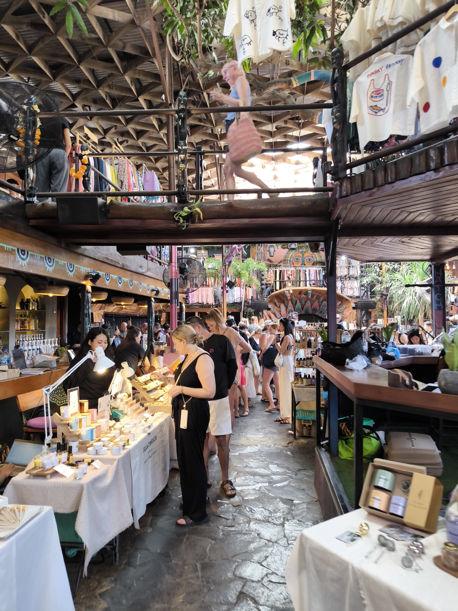 In Kuta Selatan, Indonesia, a bustling marketplace is depicted. The scene is a multi-level indoor market with a rustic, open-air feel, characterized by wooden structures, exposed beams, and lush greenery hanging from above. On the ground level, a stall displays various skincare products, including candles and bottled liquids, laid out on a white tablecloth. Several individuals are browsing or interacting with the products, including a woman in a black outfit holding a phone and a man in glasses. Further back, more stalls are visible, with racks of clothing and various items on display. The upper levels of the market are also visible, with more racks of clothing and a person walking on a walkway. The overall atmosphere suggests a vibrant and eclectic shopping environment, likely a popular tourist destination.