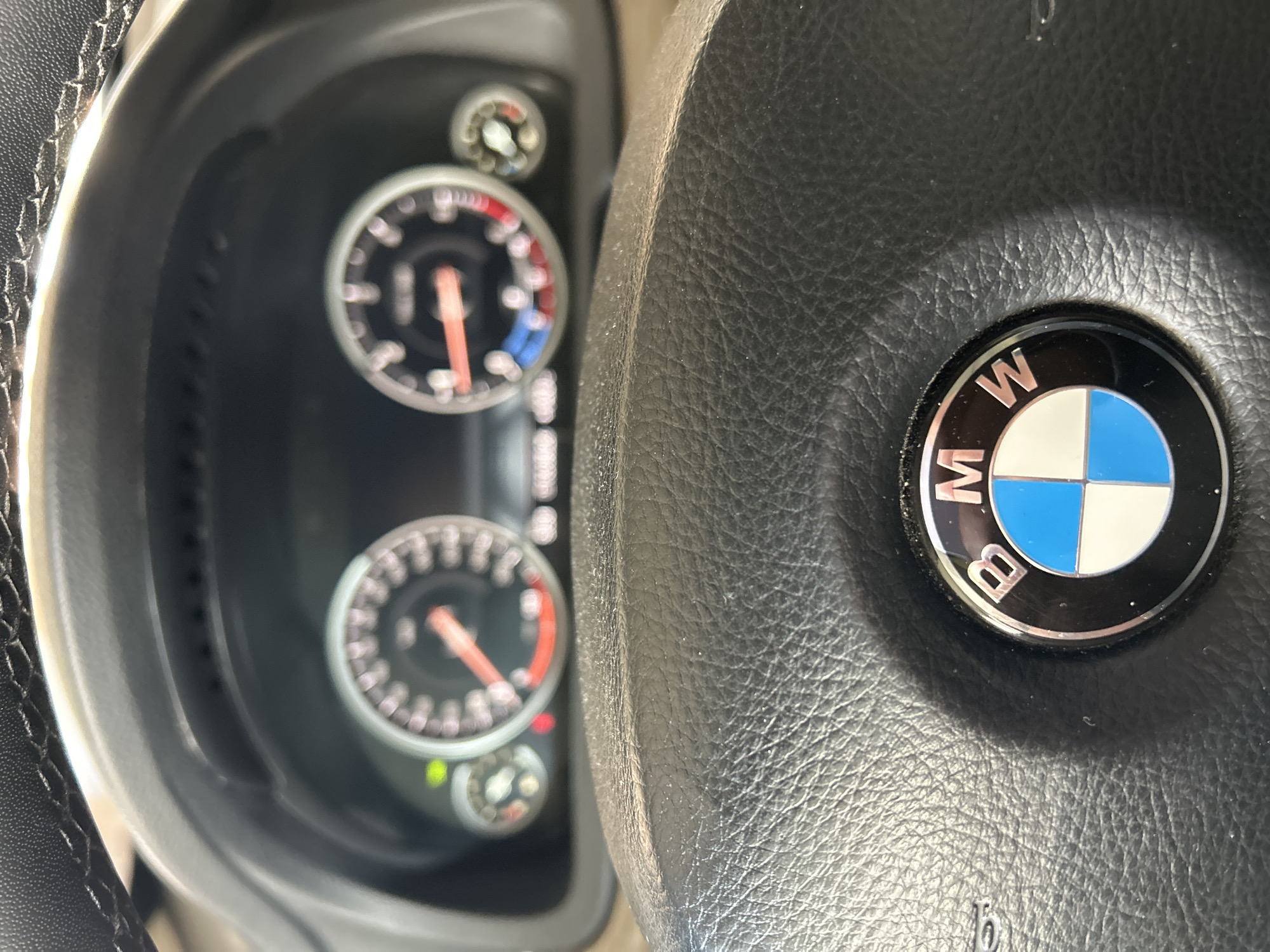 The image shows a close-up view of a BMW steering wheel and dashboard instruments. The steering wheel is made of black leather and has the BMW logo in the center, which is a round emblem with blue and white quadrants. Behind the steering wheel, the dashboard displays two prominent circular gauges, likely the speedometer and tachometer, with red needles pointing to different readings. There are also smaller indicators and lights visible on the dashboard. The lighting suggests it is daytime.