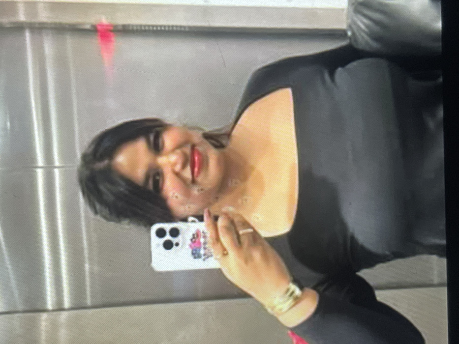 The image displays an adult female taking a selfie in an indoor environment, likely an elevator, characterized by brushed metallic wall panels. The specific location in the city of Muchiya, Nigeria, cannot be determined from the visual evidence.
The subject, appearing to be of South Asian or Middle Eastern descent, has dark hair with bangs and is smiling directly at the camera. She wears red lipstick and a long-sleeved black top with a square neckline. Her left hand holds a white smartphone, identifiable as an iPhone model with a triple camera array and a multicolored graphic case, positioned to capture the image. On her left wrist, she wears a gold-toned, wide cuff bracelet, and a ring with a light-colored stone is present on her left ring finger. The background features vertical metallic panels with a visible seam on the right side. A horizontal red light reflection is discernible in the upper right background. The lower right portion of the image shows a dark, possibly leather, object.