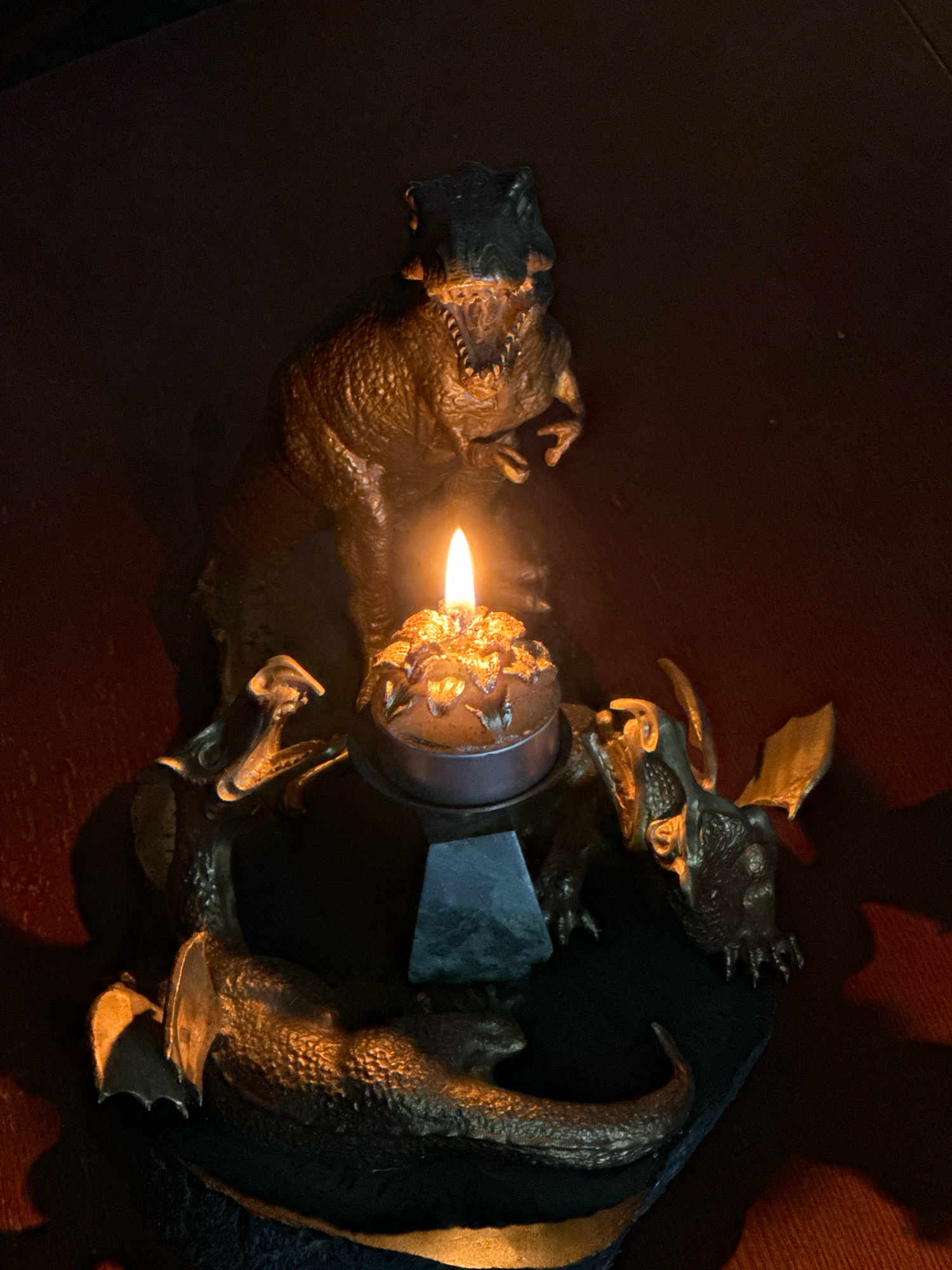 A close-up, dimly lit, eye-level shot features a candle burning in a decorative holder. The holder is adorned with at least three reptilian figures, appearing to be dragons. One dragon, larger than the others, is positioned behind the candle, with its head angled forward. Two smaller dragons are situated on either side of the candle, with their heads facing the flame. The candle itself is small and appears to be in a metallic tin, with a golden, textured wax topping. The background is dark and out of focus, with subtle reddish-brown tones. The overall impression is of a gothic or fantasy-themed decorative piece. There is no identifiable location within the city of Murmansk, Russia, as the image lacks any contextual landmarks or geographical markers.