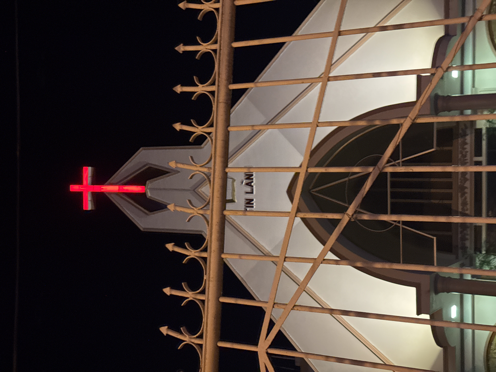 An illuminated church facade is depicted at night, viewed through a metal fence, located in Nha Trang Ward, Vietnam. The foreground features a weathered, yellowish-orange metal fence, constructed from vertical bars, horizontal supports, and diagonal bracing, augmented by decorative finials along its top edge. Beyond the fence, a brightly lit church facade, predominantly white, stands against a dark, featureless night sky. A prominent red neon or LED cross is affixed at the apex of the central gable. Below the cross, the partial text "IN LAN" is visible in dark letters on the upper white facade. The building displays a blend of angular and arched architectural elements, with subtle internal or external lighting illuminating its structure. No individuals or discernible activity are present within the scene.