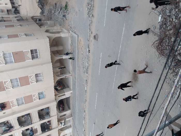 The image depicts a street scene in Khan Yunis, Palestinian Territory, captured from an elevated perspective. A multi-story, light-colored residential building with numerous windows, balconies, and an arched ground-floor arcade dominates the upper background. Laundry is visible hanging on several balconies. To the right of the building, a large pile of rubble and debris is present. In the midground, an asphalt road with a white dashed line is visible. On this road, at least seven males, primarily young adults or teenagers, are scattered, mostly clad in dark clothing, appearing to be walking or standing casually. One individual in the lower center wears a light brown shirt. In the building's arcade, several figures, including adults and a child, are seen. A red vehicle is parked beneath the arcade. The immediate foreground features multiple utility wires and a substantial pile of tangled rebar and concrete debris.