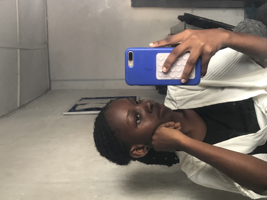 The image displays a young adult female of African descent taking a mirror selfie indoors. She has dark, braided hair pulled back and wears a black top beneath a light-colored, pleated wrap or cardigan. A black, four-leaf clover-shaped stud earring is present in her left ear. Her left hand is positioned under her chin, while her right hand holds a smartphone with a blue case vertically. The phone case features a visible Apple logo and a white rectangular attachment with a grid of circular elements on its back. The background, visible in the mirror's reflection, consists of a light gray, textured wall and a light-colored ceiling with discernible seams. A dark-framed object containing a blue element is partially visible on the wall behind the subject's left side. An indistinct dark object is reflected behind her right shoulder. The specific location within the city of Ado, Nigeria, is not visually identifiable from the image; however, the scene depicts an interior room.