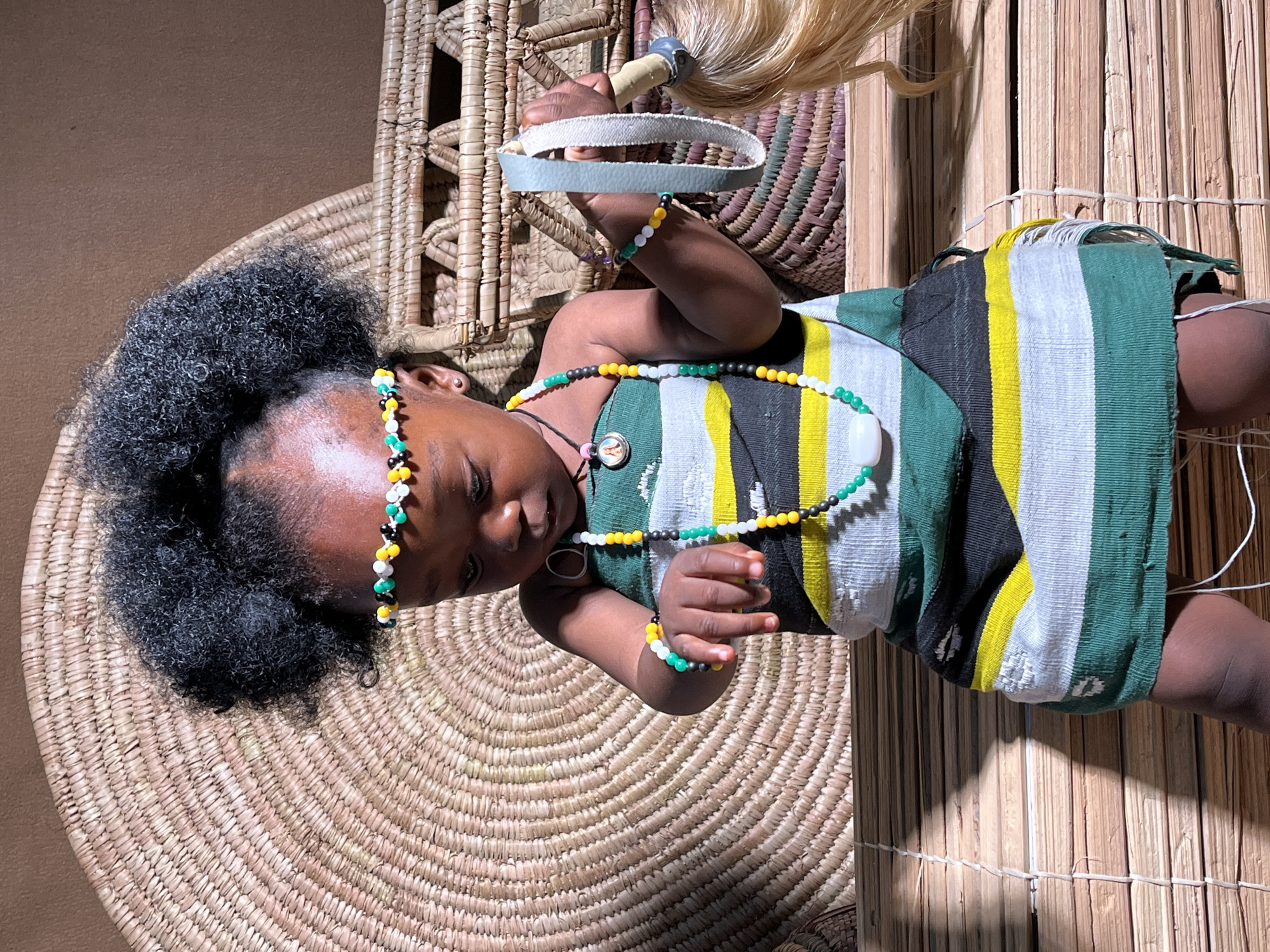 The image shows a child lying on a woven mat. The child has dark skin, dark curly hair and is wearing a beaded headpiece and necklace. The child is wearing a striped dress of green, yellow, black and white. The child is holding a toy with a white handle and a blonde fringe. The background consists of woven baskets and mats and wooden panels. The scene is likely indoors. The image contains no specific details to place it in Barakin Akawo, Nigeria.
