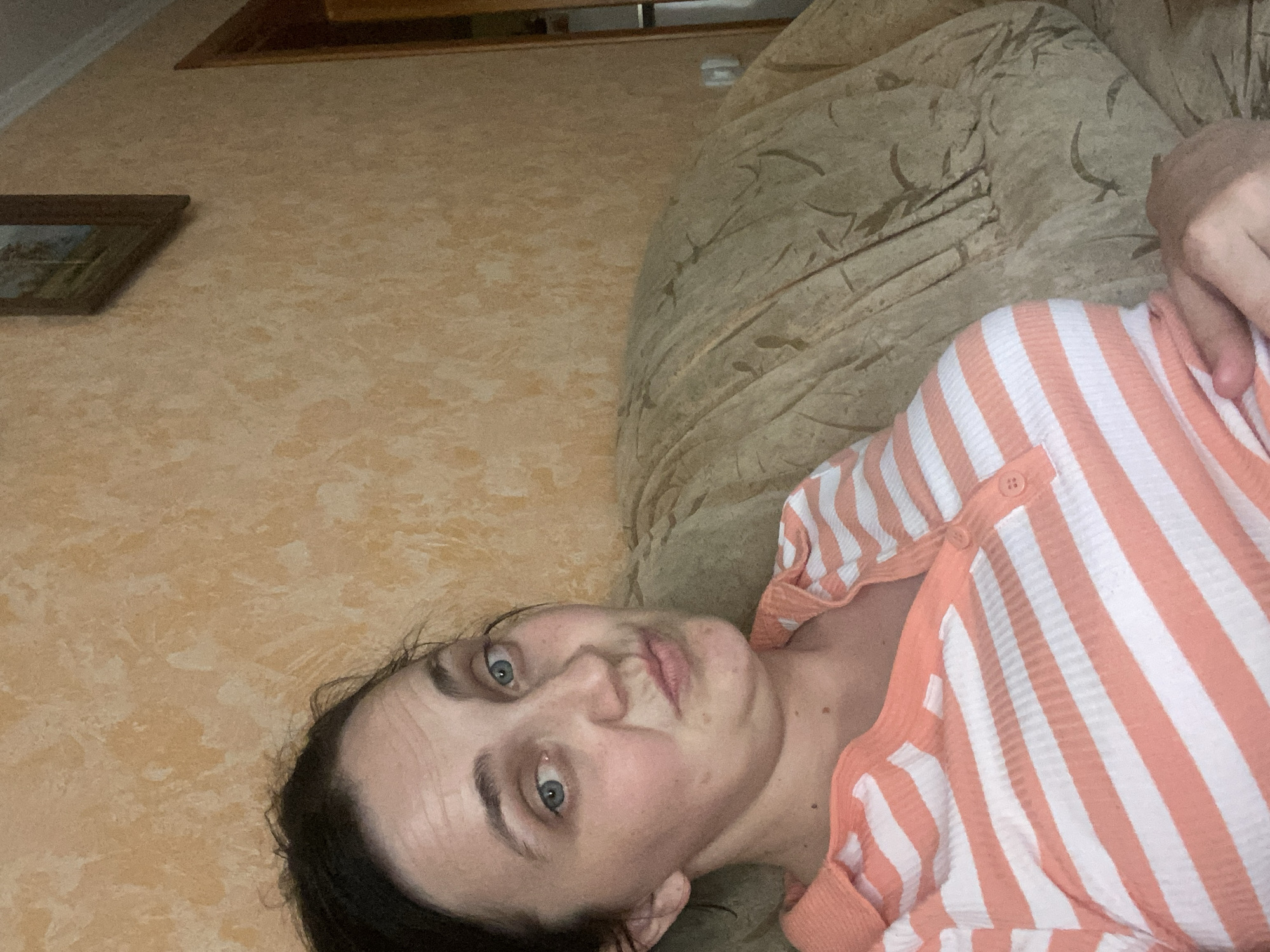 The image shows a young woman lying on a couch, looking at the camera. She has dark hair and blue eyes, and is wearing a striped shirt with pink and white stripes. The couch appears to be made of a textured fabric, possibly a sectional sofa, and is a neutral beige color with a subtle pattern. Behind the couch, there is a wall with a light peach or tan textured wallpaper. A framed picture hangs on the wall to the left of the couch. The lighting in the room appears to be artificial, suggesting it could be evening or a dimly lit interior. The woman's expression is neutral, with a slight pout to her lips. The overall scene suggests a casual, relaxed moment at home. There is no visible text in the image.