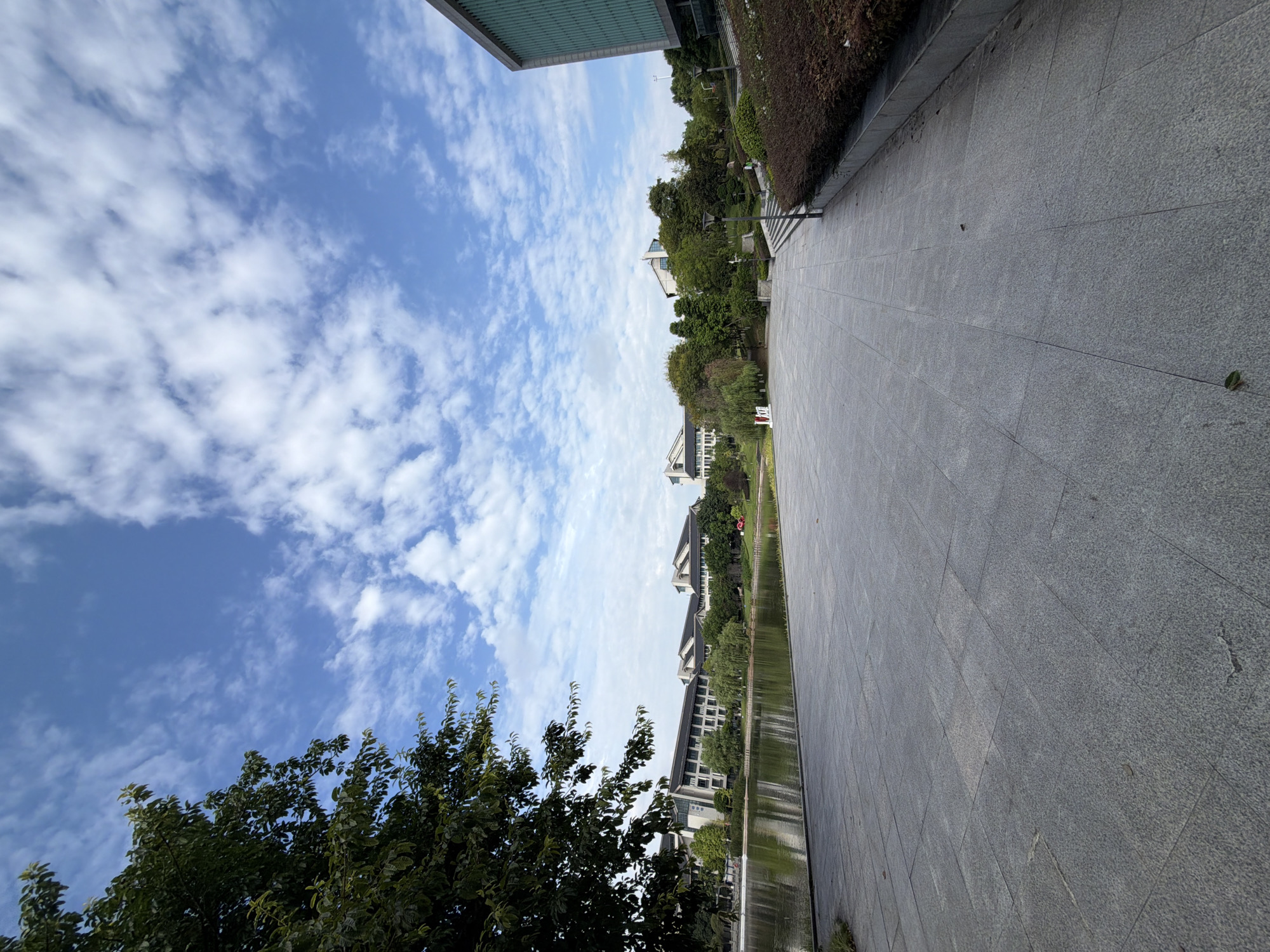 The image depicts a wide, paved plaza area in front of a body of water, likely a lake or pond, with buildings and lush greenery in the background. The sky is a vibrant blue, populated with scattered white clouds. On the left side of the frame, a large tree with dark green foliage partially obscures the view of several low-rise buildings with dark roofs. The buildings appear to be part of an institutional complex, possibly a university or government facility.
The plaza itself is made of large, grey, rectangular stone tiles. To the right of the plaza, there is a sloped area with neatly trimmed brown bushes and some smaller trees, leading up to a modern building with a teal-green facade. Several lampposts are visible along the pathway. The water reflects the sky and the surrounding trees. The overall scene suggests a well-maintained public space on a clear day. The location is identified as Jiangjin, China. There are no people visible in the image, and no discernable actions or interactions are taking place.