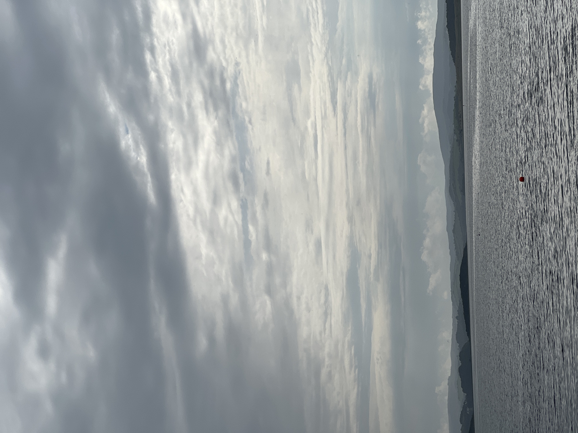 The image shows a wide shot of a body of water, likely a bay or ocean, with a hilly coastline in the distance under a cloudy sky. The water is dark and appears choppy. The coastline is a series of rolling hills with varying shades of green and gray. The sky is dominated by layers of gray clouds with some lighter, brighter patches indicating potential sunlight breaking through. In the foreground, a single red buoy is visible on the surface of the water. The scene appears to be a natural landscape, possibly a coastal area. The location is identified as Mountcharles, Ireland, suggesting this is a view of the Donegal Bay area. There are no people or discernable actions visible in the image, making it a still, environmental shot.