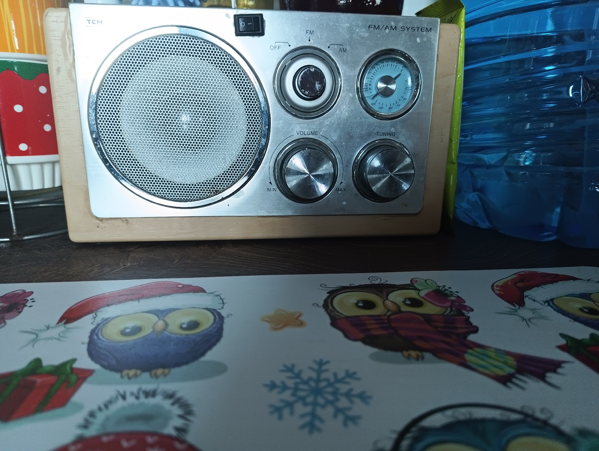 The image depicts a close-up view of a silver TCM FM/AM radio positioned on a wooden surface. The radio has a prominent speaker grill on the left, a power switch at the top, and several knobs and a tuning dial. To the right of the radio is a large, clear blue plastic water jug with visible internal plastic structures. In the foreground, a white paper or placemat features cartoonish illustrations of owls wearing Santa hats and scarves, alongside festive elements like snowflakes and wrapped gifts, suggesting a holiday theme. The setting appears to be indoors, likely a domestic environment, with no discernible elements that would specifically place it in Karnobat, Bulgaria.