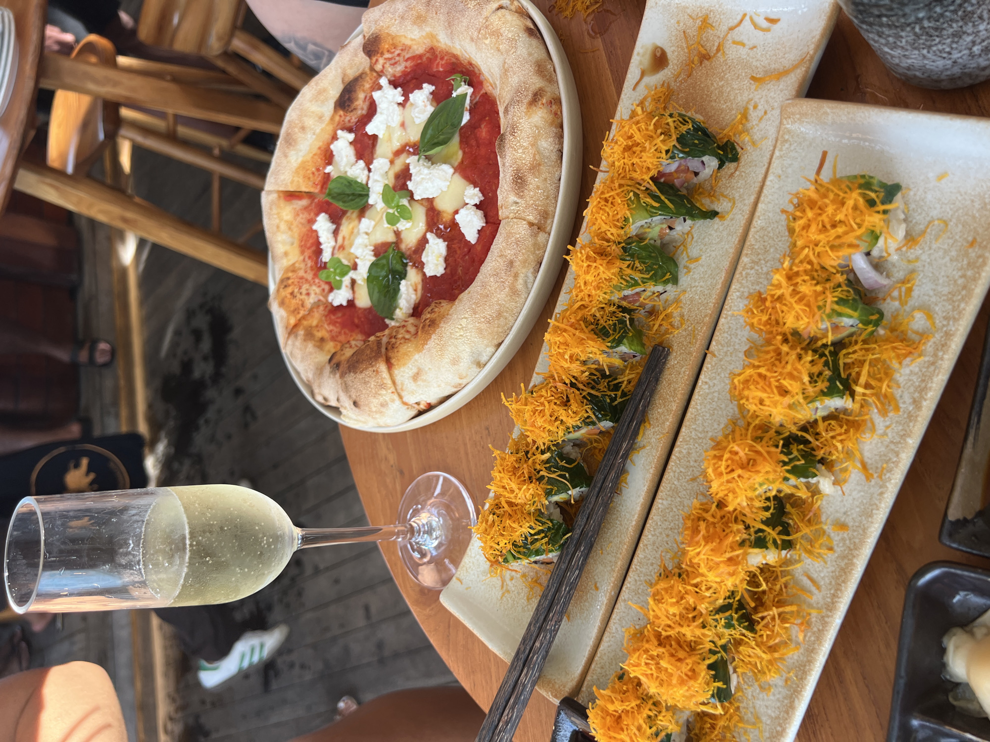 In Ratsada, Thailand, a wooden table displays a Margherita pizza topped with tomato sauce, fresh mozzarella, and basil. Beside the pizza, two long plates hold sushi rolls; the rolls are wrapped in what appears to be green leaves and topped with shredded fried yellow noodles. A flute glass filled with a bubbly, light-colored beverage, likely champagne, stands to the left of the pizza. Chopsticks rest on the sushi plates. In the background, blurred figures and wooden chairs suggest a restaurant setting. The floor is made of dark wood planks.