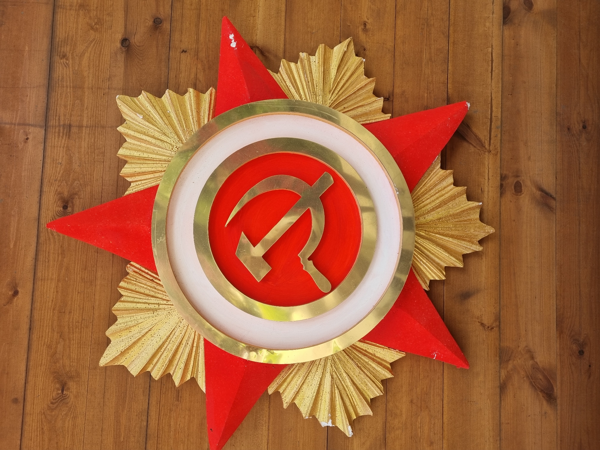 The image shows a wooden plaque featuring the Soviet hammer and sickle emblem. The emblem is centrally positioned within a concentric ring, painted gold and white, against a red background. The plaque is shaped like an eight-pointed star, with alternating red and gold-colored points. The plaque is mounted on a brown wooden wall. The location is not specified in the image.