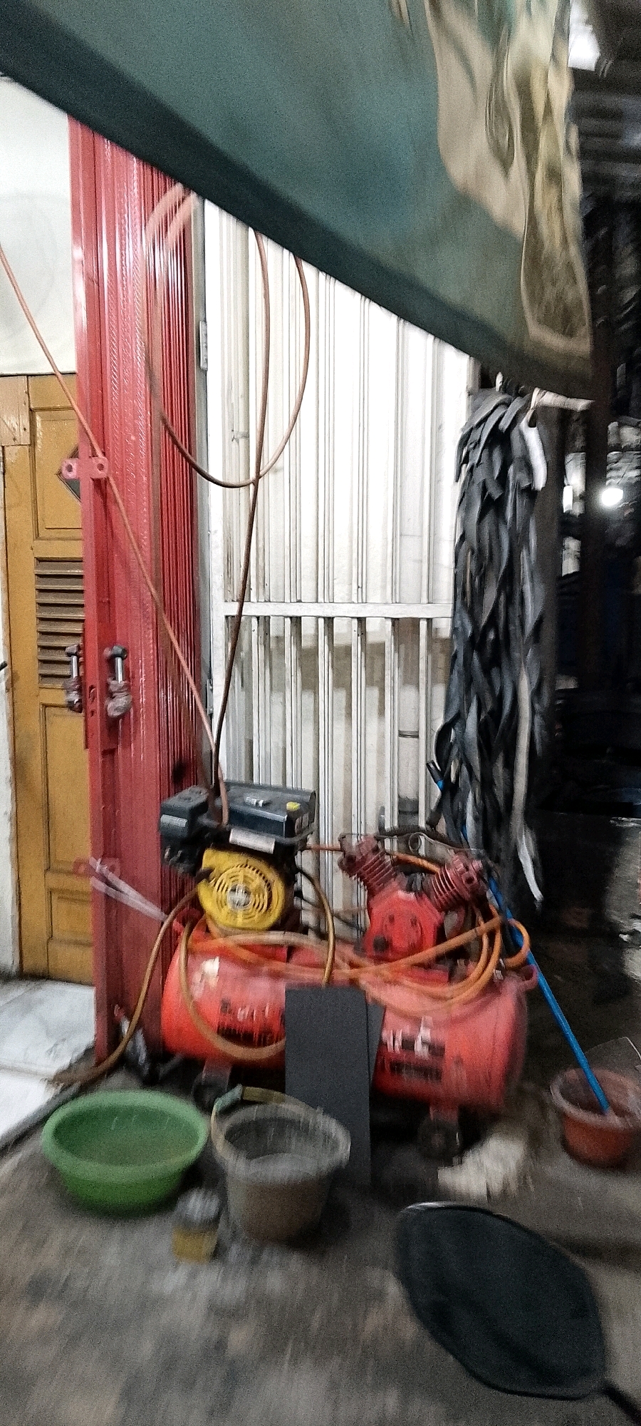 In Depok, Indonesia, a red and yellow air compressor with connected hoses is situated on a gray concrete floor in front of a light-colored corrugated metal roll-up door. To the left, a red metal pole with visible bolts and a yellow wooden door with louvers are present. In the foreground, a green plastic basin, a small brown bucket filled with a gritty substance, and a dark gray rectangular object are positioned on the floor. To the right, a collection of dark, layered material, resembling cut tires, hangs from a horizontal support, and further right, a red plastic bowl containing a blue object is visible. The scene is dimly lit, with a dark green awning or ceiling element overhead reflecting a light-colored, abstract image.