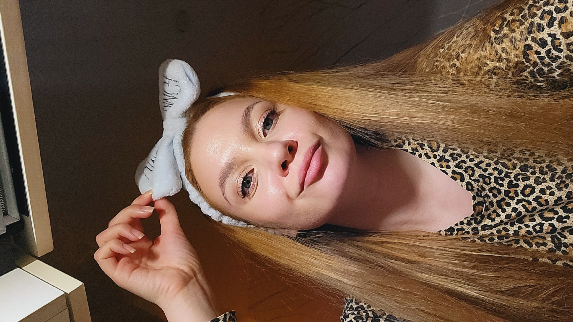 A young woman with blonde hair and blue eyes, wearing a blue headband and a leopard-print top, poses in a dimly lit room. Her face is tilted towards the camera with a slight smile. Her right hand is near the headband. A piece of furniture is visible on the left. The photograph was taken in Chaikovsky, Russia.