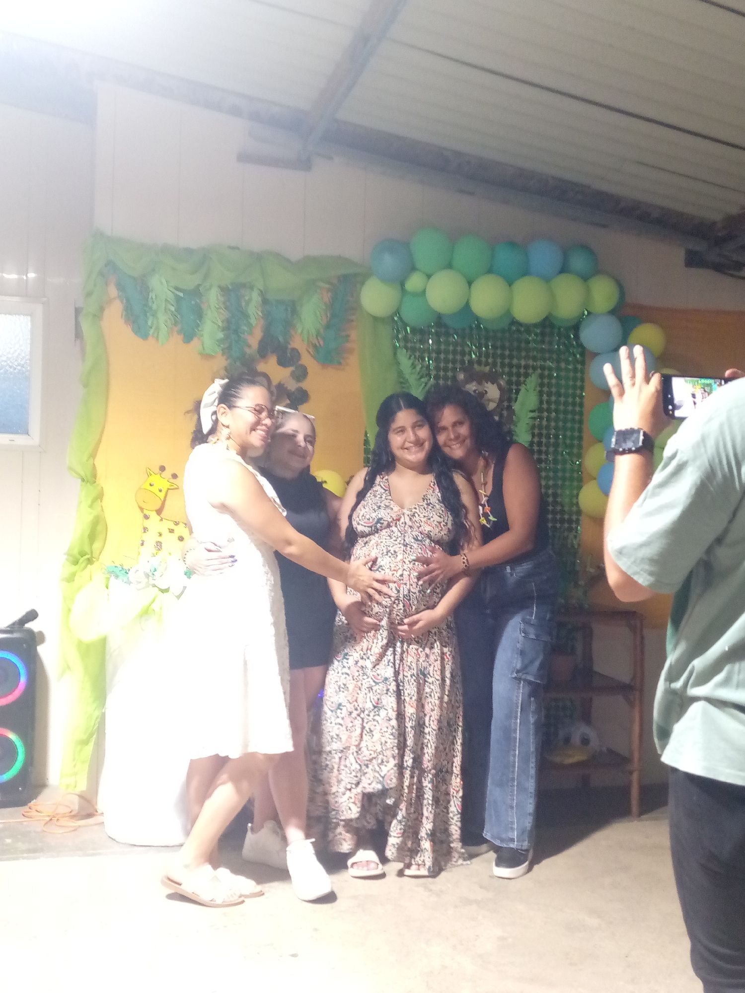 The image shows a group of women posing at what appears to be a baby shower in Altagracia, Venezuela. A pregnant woman in a long, patterned dress is at the center, with others placing their hands on her stomach. Behind them is a decorated wall with a yellow backdrop, green leafy decorations, and blue and green balloons. To the left is a speaker with colorful lights. A person on the right is taking a photo with a smartphone.
