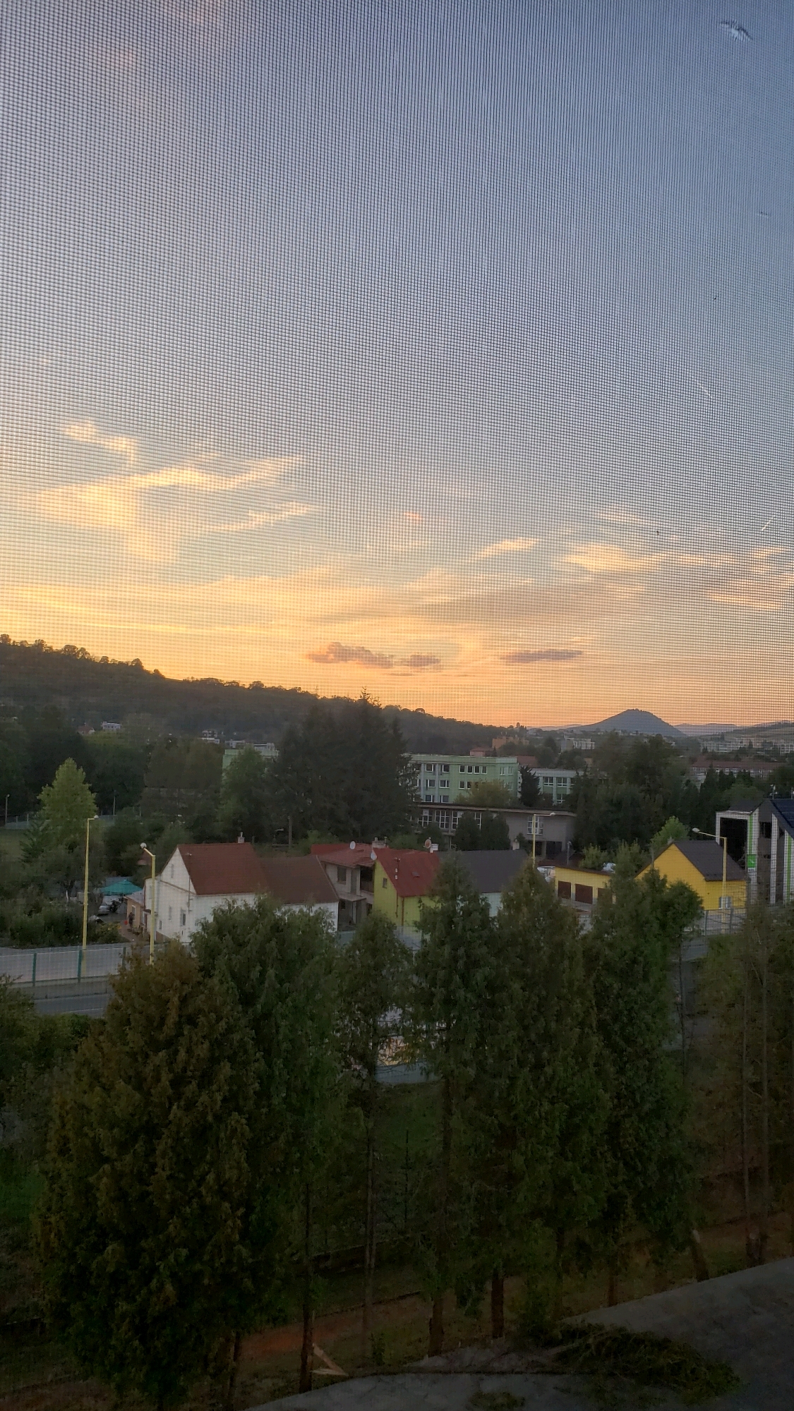 The image depicts a sunset over a residential area, likely in Prešov, Slovakia, viewed through a window screen. In the foreground, several evergreen trees are visible, some tall and slender, others bushier. Behind the trees, a street runs horizontally, bordered by a fence. Several houses with red and orange tiled roofs are situated along the street, some appearing to be under construction or renovation. In the middle ground, a row of green apartment buildings stands out, with a larger, lighter green building further back. Beyond the buildings, rolling hills covered in trees recede into the distance, culminating in a prominent, rounded mountain peak against the sunset sky. The sky itself is a gradient of pale blue at the top, transitioning to soft oranges, yellows, and pinks closer to the horizon, with wispy clouds scattered throughout. The overall scene suggests a peaceful, suburban evening in Prešov.