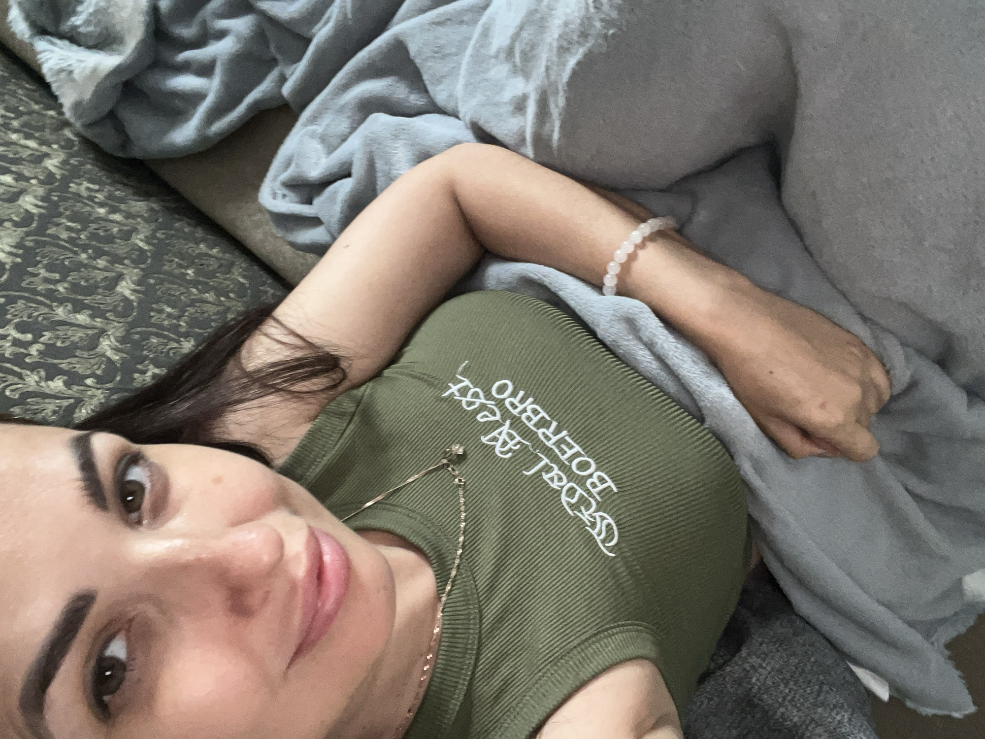 The image is a close-up, high-angle selfie of a woman lying down, partially covered by blankets. She has dark hair and is looking directly at the camera. Her expression is relaxed, with a slight smile. She is wearing a green, ribbed tank top with white text embroidered on it. The text appears to be "SKAL BEST BOERBRO". She is also wearing a delicate gold necklace and a pink beaded bracelet on her left wrist. The background consists of a patterned sofa and soft, grey blankets, suggesting a comfortable indoor setting, possibly a bedroom or living room. The lighting is soft, indicating it could be daytime or evening. The overall mood is intimate and casual.