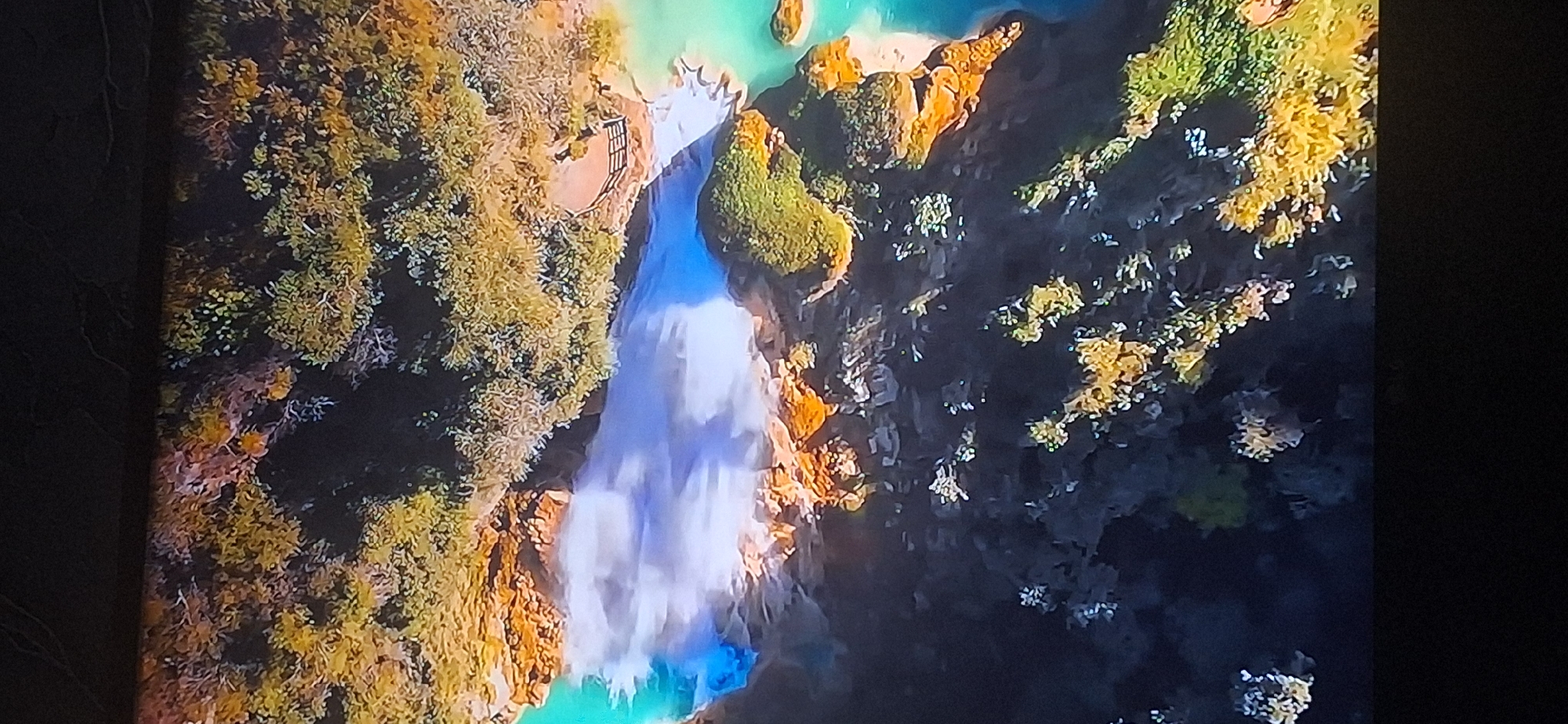 This is an aerial drone shot of a natural landscape featuring a waterfall with turquoise water cascading over orange rock formations. Lush green and yellow vegetation covers the surrounding cliffs and terrain. A wooden walkway with railings is visible on one of the cliff edges, overlooking the waterfall. The scene appears to be set in a natural park or a scenic wilderness area. There are no people or man-made structures other than the walkway visible in the frame. The location is not in Bila Tserkva, Ukraine.