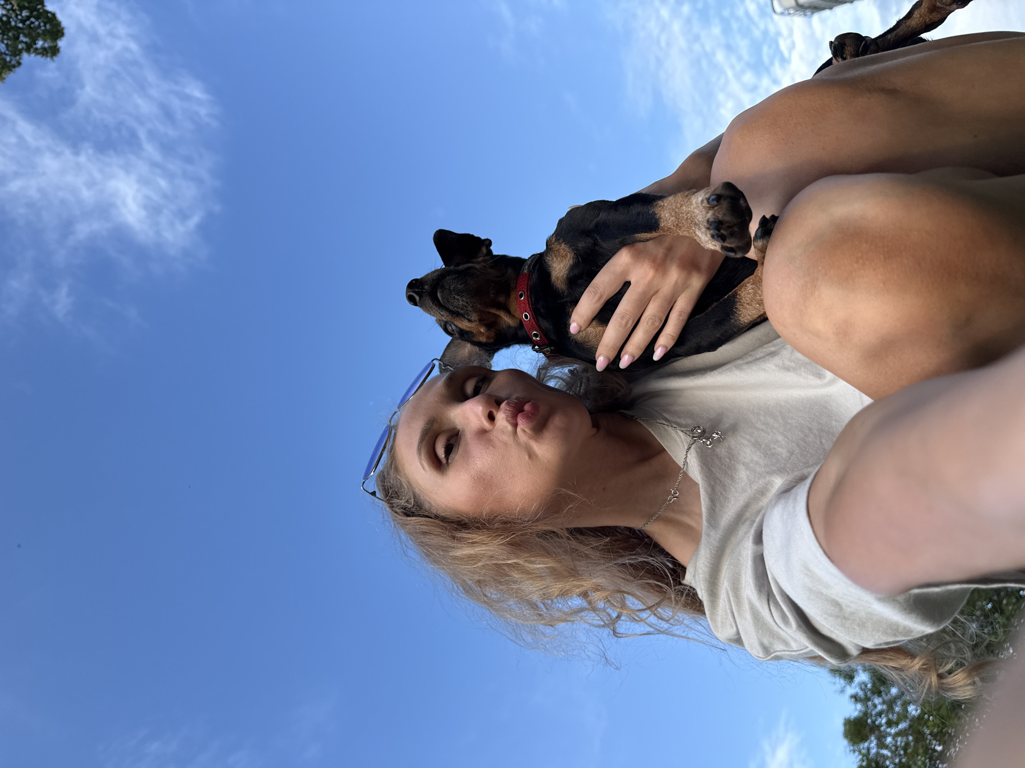 An outdoor, low-angle, eye-level, close-up, medium shot shows a young woman with blonde, curly hair and a light-colored t-shirt holding a small, black and brown dog with a red collar. The woman is puckering her lips for a selfie and looking towards the camera. Her left hand is holding the dog's body while the dog looks up towards the sky. The background is a bright blue sky with wispy clouds. To the left of the frame, the top of some trees can be seen. No specific location in Rosstani, Russia is identifiable in the image.