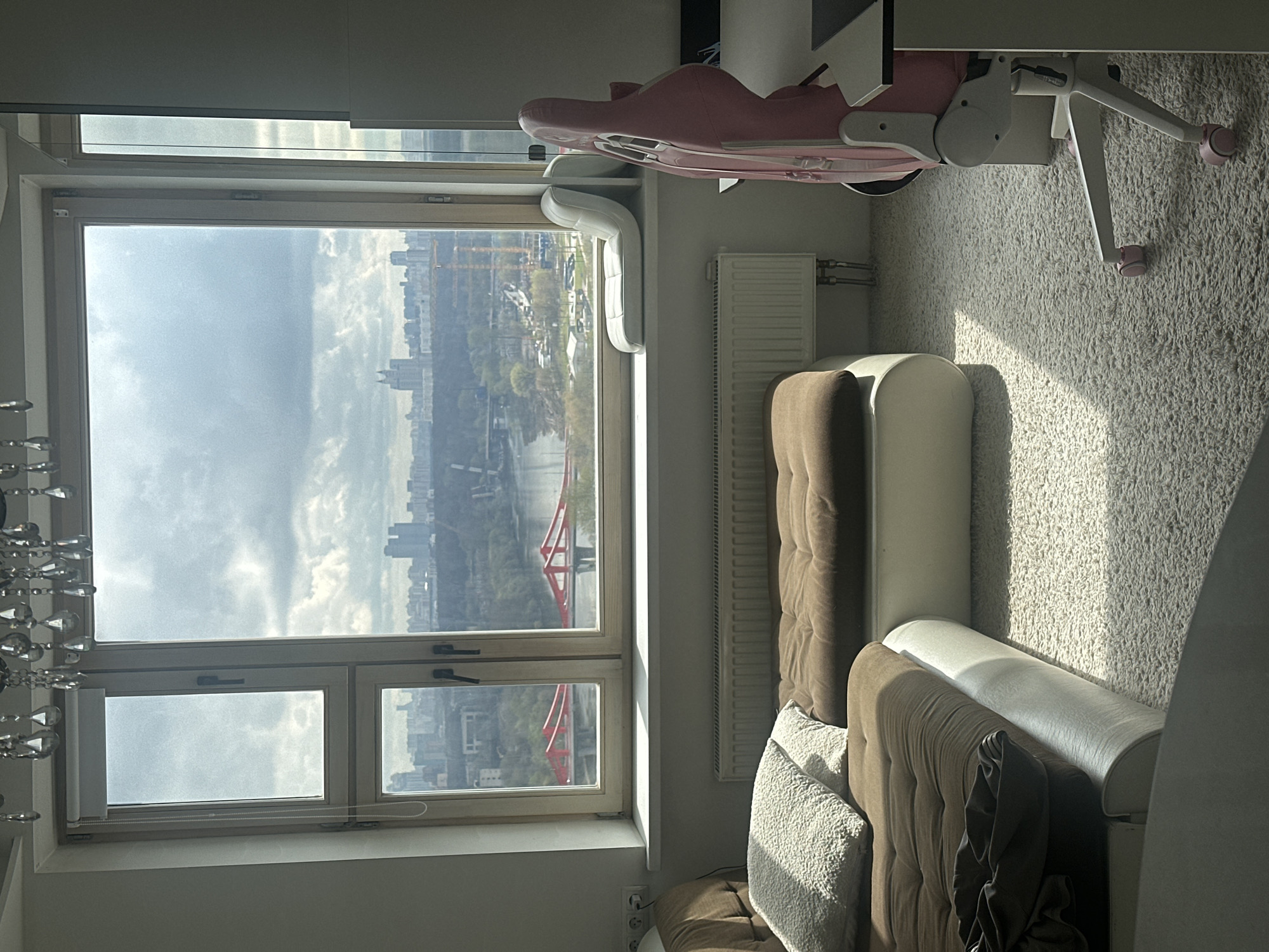 The image shows a bright indoor room with a large window offering a panoramic view of an urban landscape. The room features a light-colored, shaggy carpet covering the floor. Sunlight streams through the window, casting distinct shadows across the carpet and furniture, indicating it is daytime, likely late morning or early afternoon, under a partly cloudy sky.
To the right of the window, a white radiator is mounted below the windowsill. Further into the room, two sections of what appear to be sofas or armchairs are visible, upholstered in a brown, tufted fabric with lighter-colored bases. One armchair includes a textured white cushion. In the upper right corner, a pink and white ergonomic chair with wheels is partially seen, possibly part of a desk setup. On the far left, a decorative light fixture with multiple clear, faceted elements hangs. A white roller blind is partially drawn on one of the lower window panes. Electrical outlets are visible near the floor next to the furniture.
Through the multi-pane window, a city skyline extends into the distance. A body of water, likely a river, flows through the urban area, crossed by a prominent red bridge with a distinctive structure. Green trees line the riverbanks, and various city buildings, some with visible construction cranes, form the urban vista. No people or specific activities are depicted. The architectural and landscape features are consistent with the provided location context of Khimki, Russia. There is no visible text.