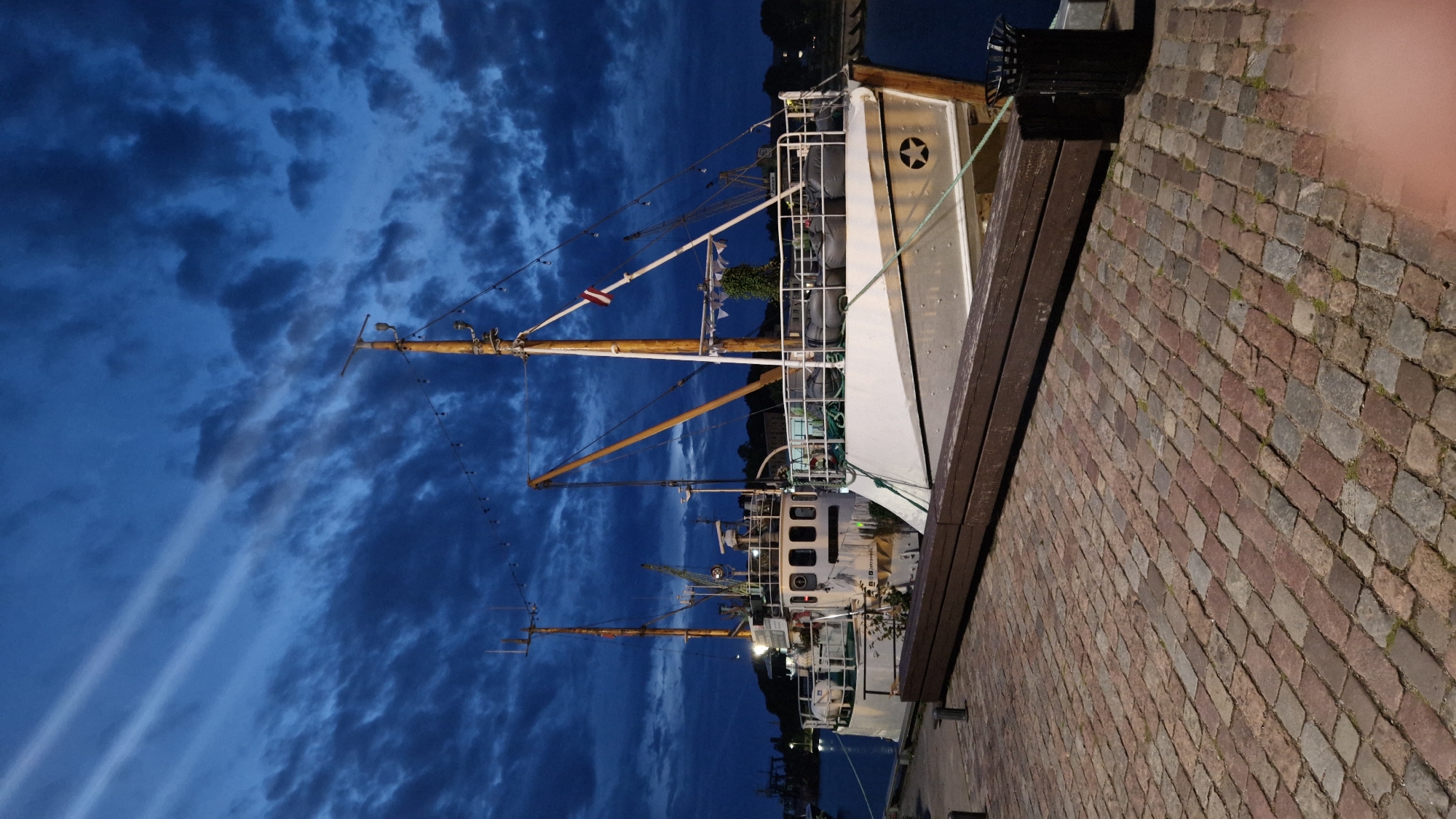 The image depicts two docked boats at dusk in Liepaja, Latvia. The boats are white with wooden accents and rigging, and are tied to a cobblestone quay. The sky is dark blue with dramatic clouds, illuminated by some artificial lights. A wooden bench runs along the quay in the foreground, and a trash can is visible on the right. The scene is calm and appears to be a quiet harbor setting.